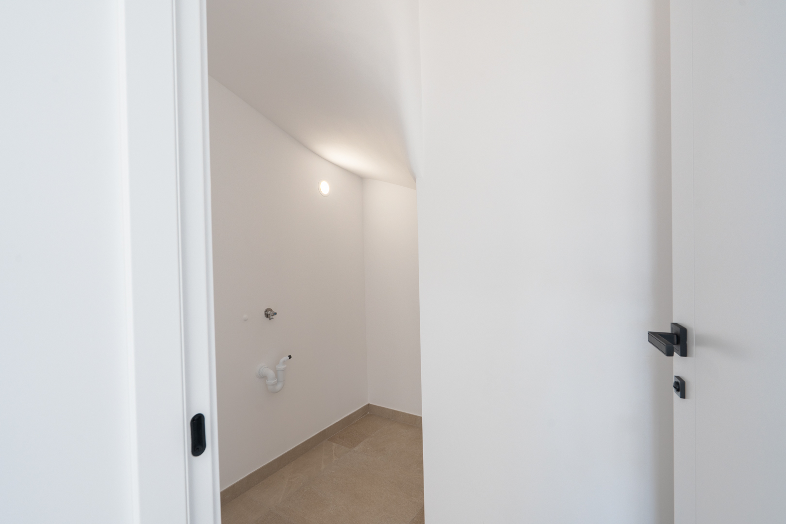 Practical utility or storage room featuring white walls, porcelain flooring, and pre-installed plumbing connections under a sloped ceiling.