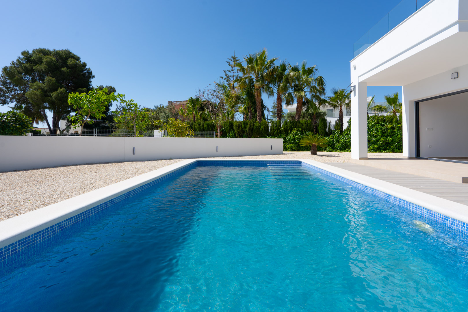 Rectangular swimming pool with blue mosaic tiling, bordered by white gravel and modern perimeter walls featuring integrated lighting and palm trees.