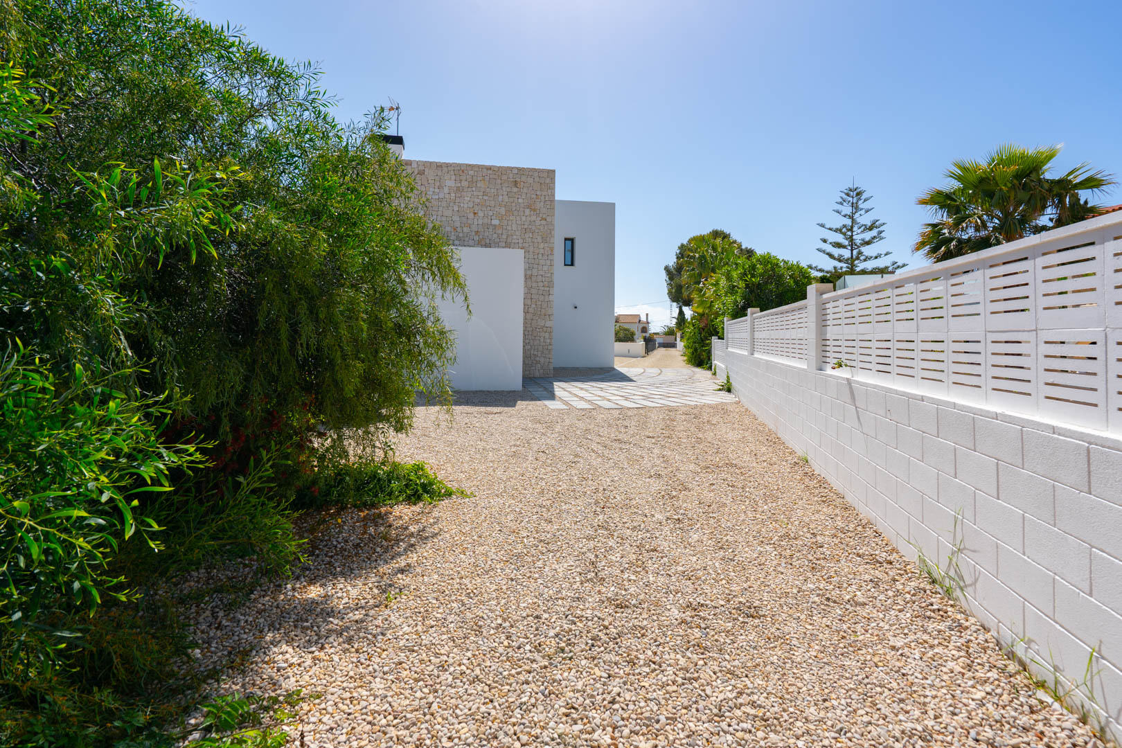 Side entrance featuring a gravel driveway, white perimeter wall, and a facade blending natural stone cladding with modern white finishes.