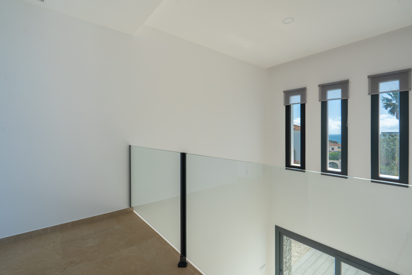 Upper floor landing featuring a glass balustrade overlooking the double-height void. Three vertical windows with grey blinds provide sea views and natural light.