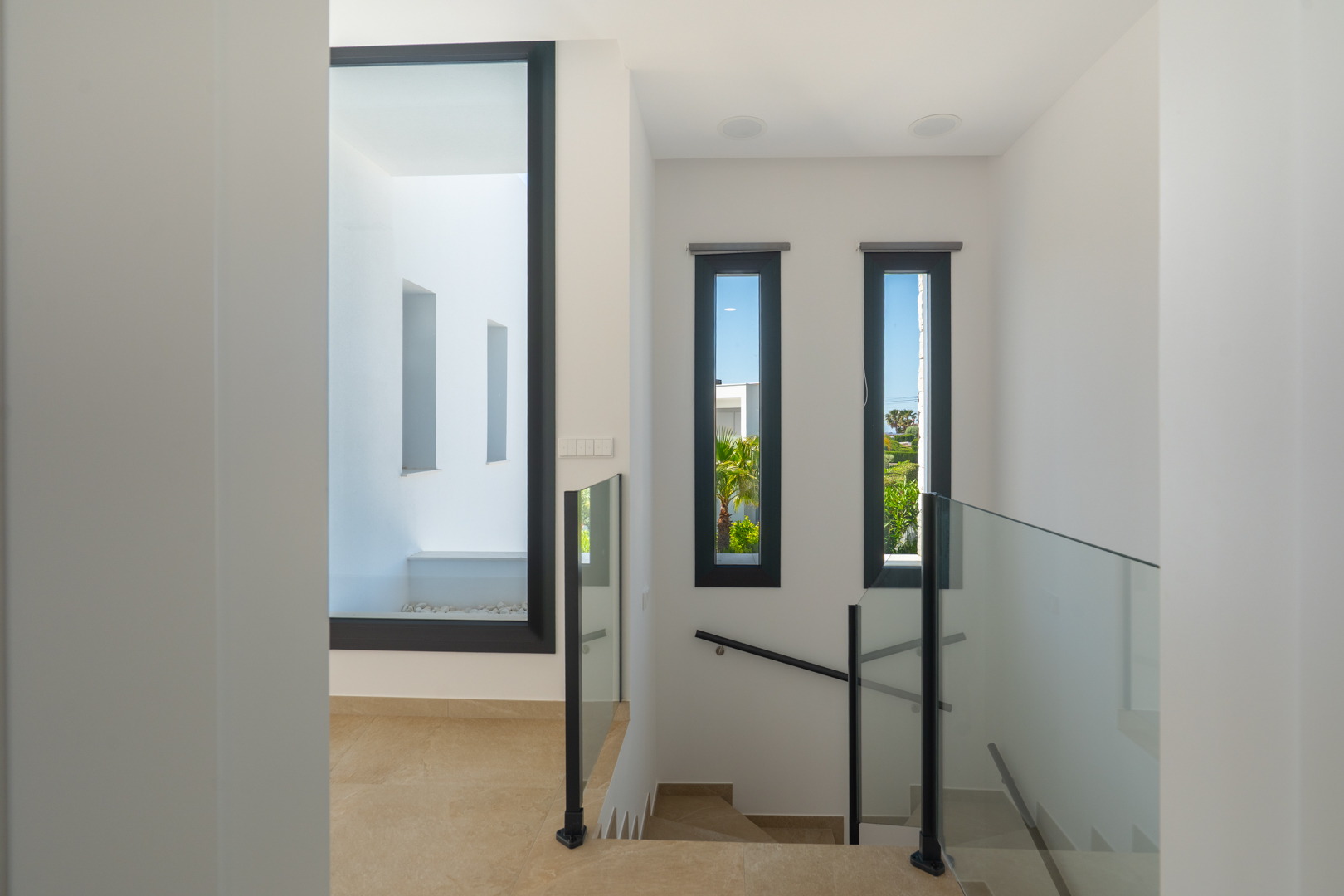 Upper floor landing featuring glass balustrades, porcelain flooring, and large vertical windows providing natural light and views of the surrounding greenery.