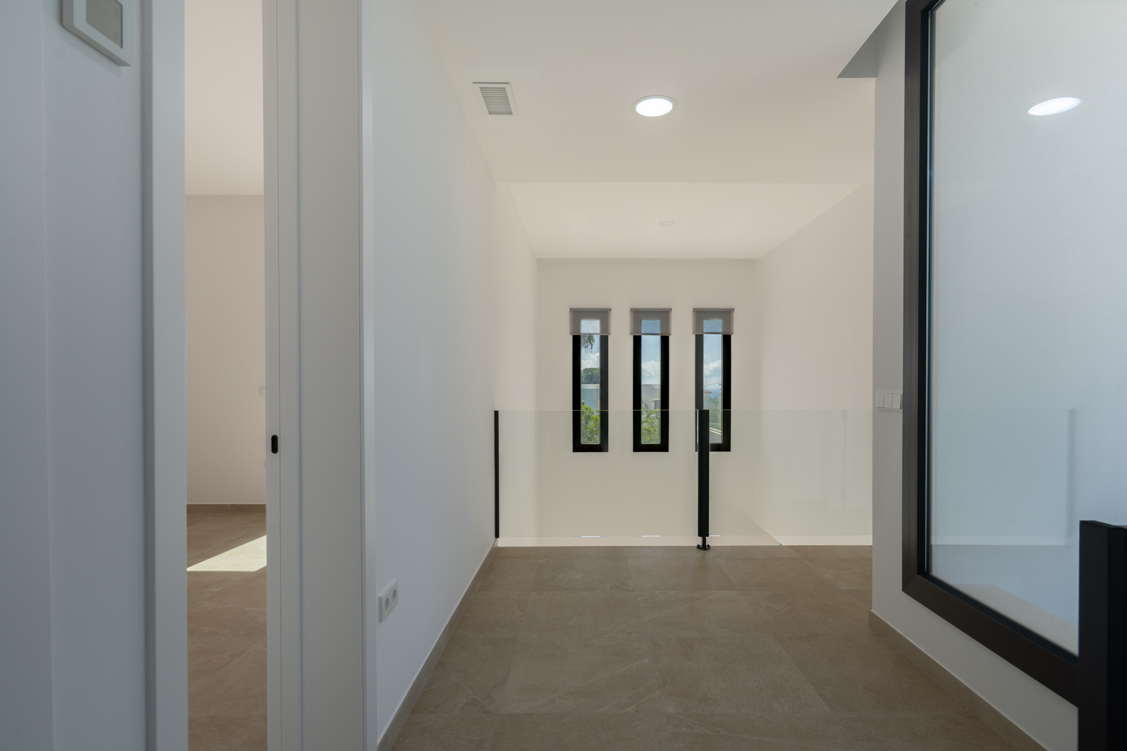 Upper floor landing featuring porcelain tile flooring, a glass balustrade, and three narrow vertical windows overlooking the double-height living area.