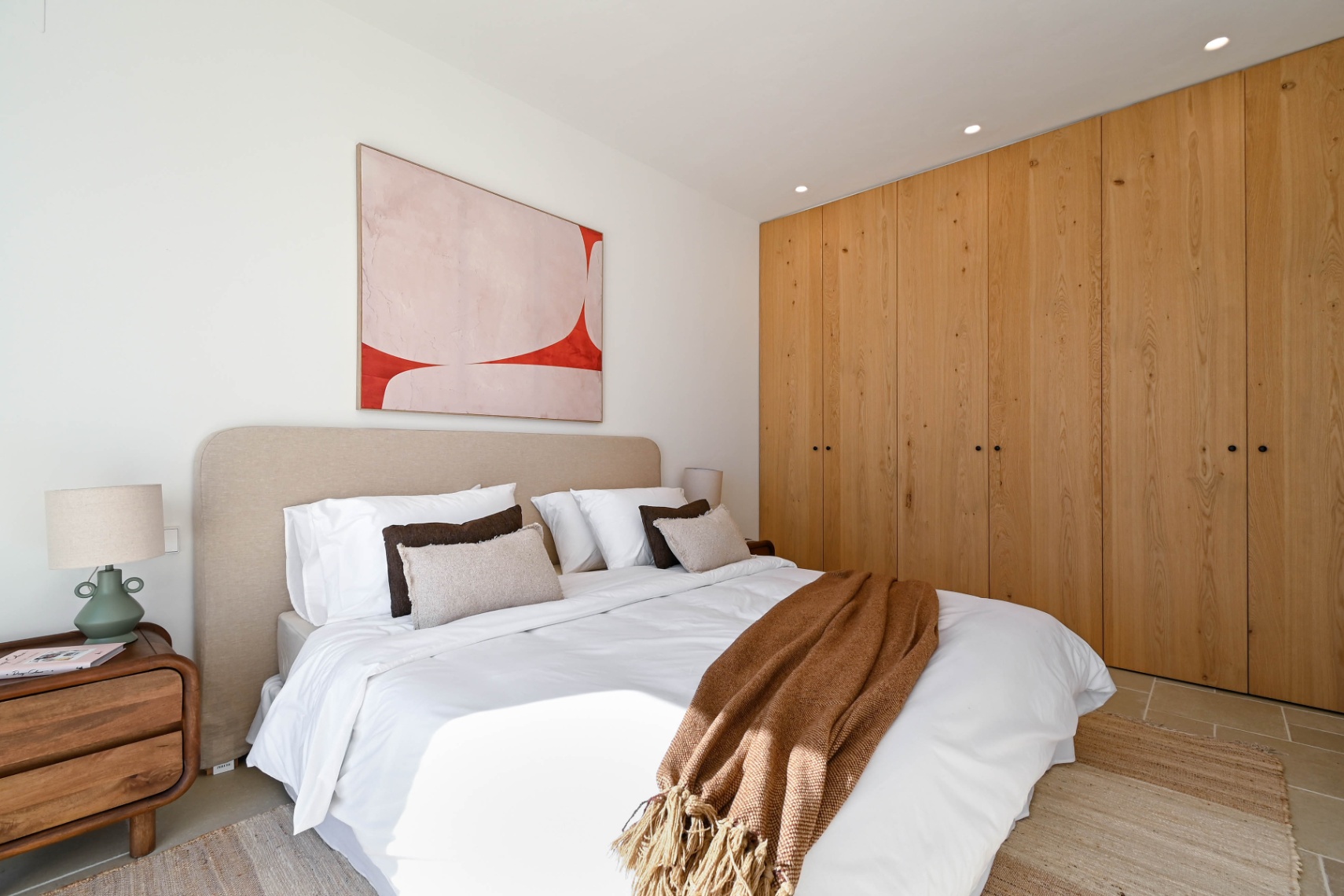 Bedroom featuring floor-to-ceiling natural wood wardrobes, a beige upholstered headboard, and recessed ceiling lighting. The space includes tiled flooring and minimalist abstract art.