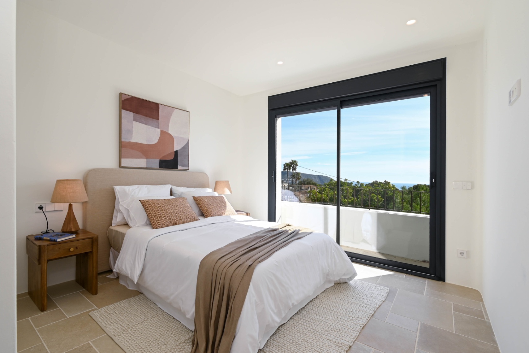 Bright bedroom featuring natural stone flooring and large sliding glass doors opening onto a private terrace with views of the Mediterranean landscape.