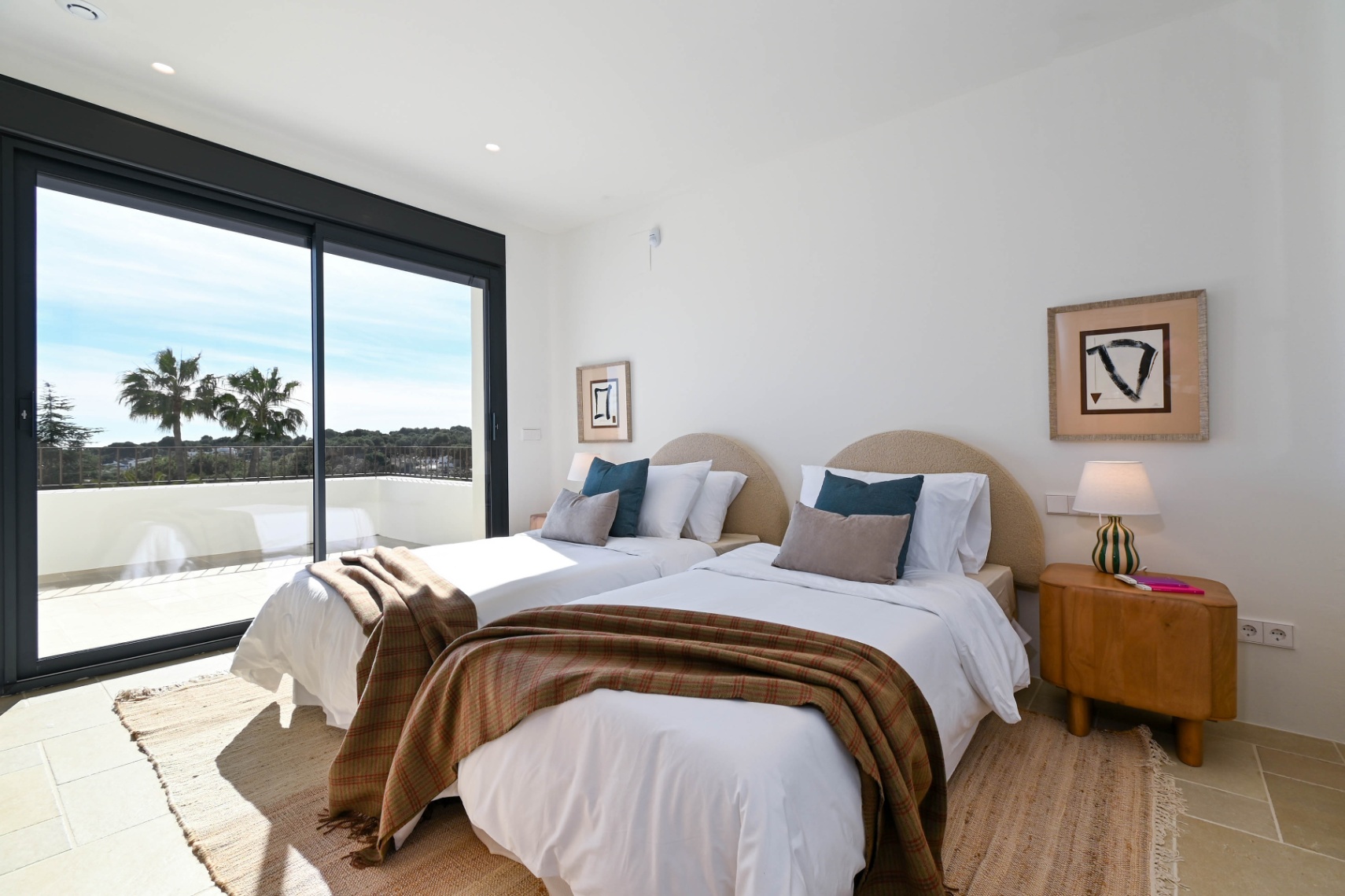Bright bedroom featuring twin beds with curved headboards and direct access via sliding glass doors to a terrace with views of local greenery and palms.