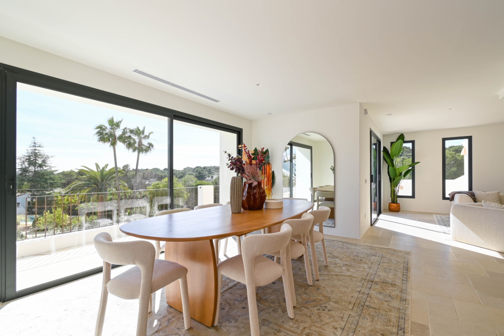 Bright dining area featuring an oval wooden table and cream chairs, with large sliding doors opening to a terrace with views of palm trees and Mediterranean greenery.
