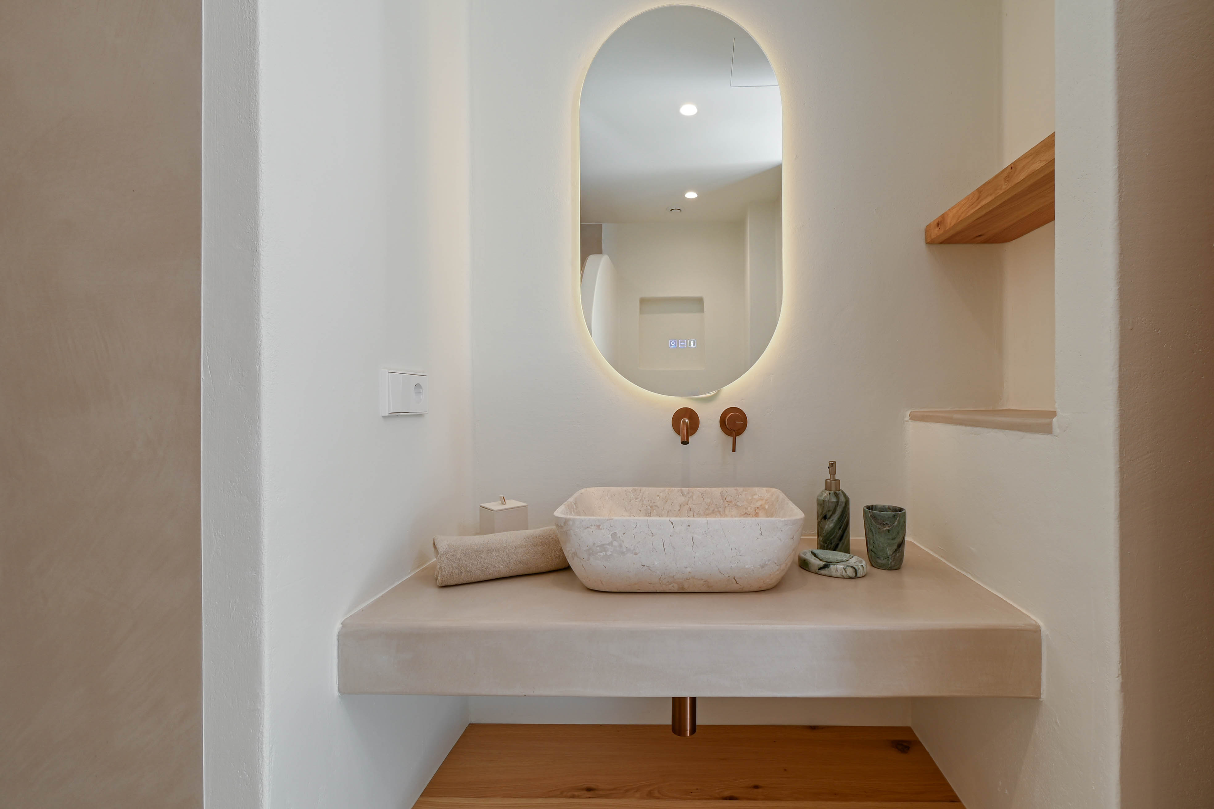 Designer bathroom featuring a natural stone vessel sink on a microcement vanity, copper wall-mounted faucets, and an oval backlit mirror with oak accents.