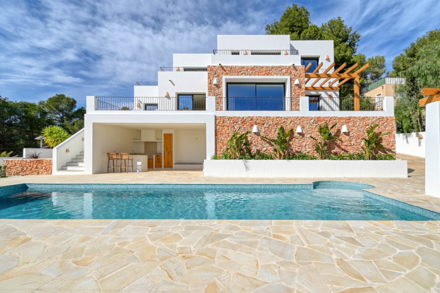 Exterior view featuring a blue mosaic pool, natural stone terrace, and a multi-level facade with dry stone walls, a covered outdoor kitchen, and a wooden pergola.