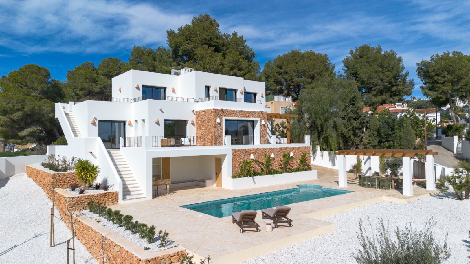 Exterior view of the modern villa featuring white facades, natural stone walls, a rectangular swimming pool, and multiple terraces with Mediterranean landscaping.