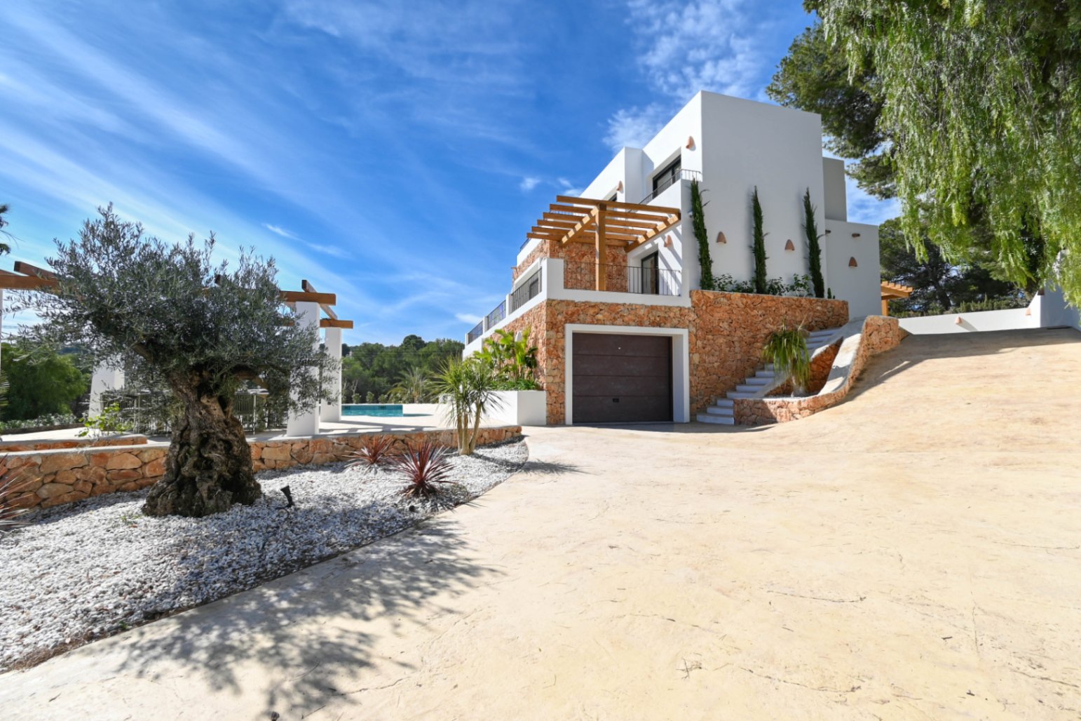 Exterior view of the villa featuring natural stone cladding, an integrated garage, and a paved driveway bordered by an olive tree and Mediterranean landscaping.