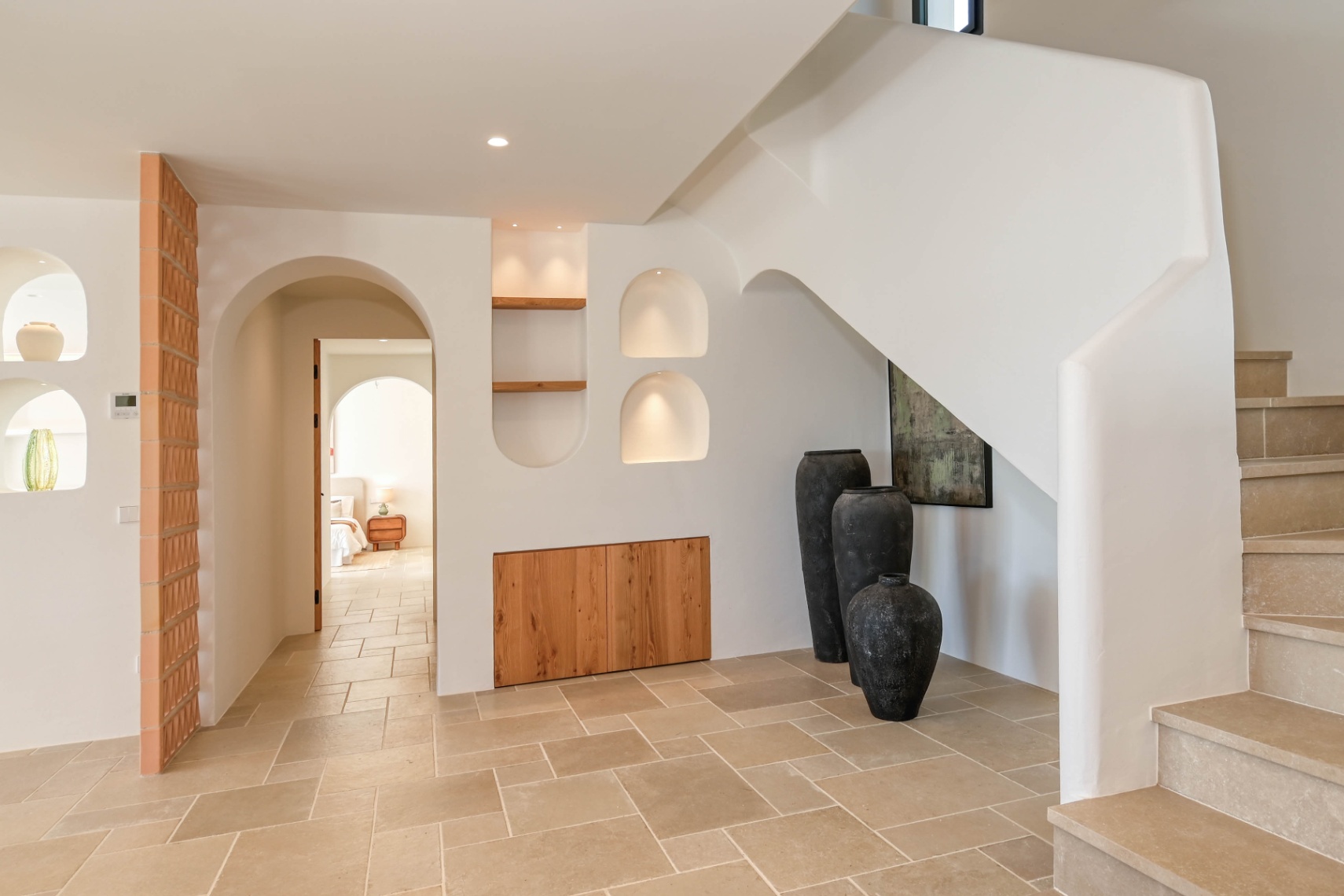 Mediterranean-style hallway featuring a stone staircase, illuminated wall niches with wood shelving, and integrated cabinetry. Natural stone flooring flows through elegant arches.