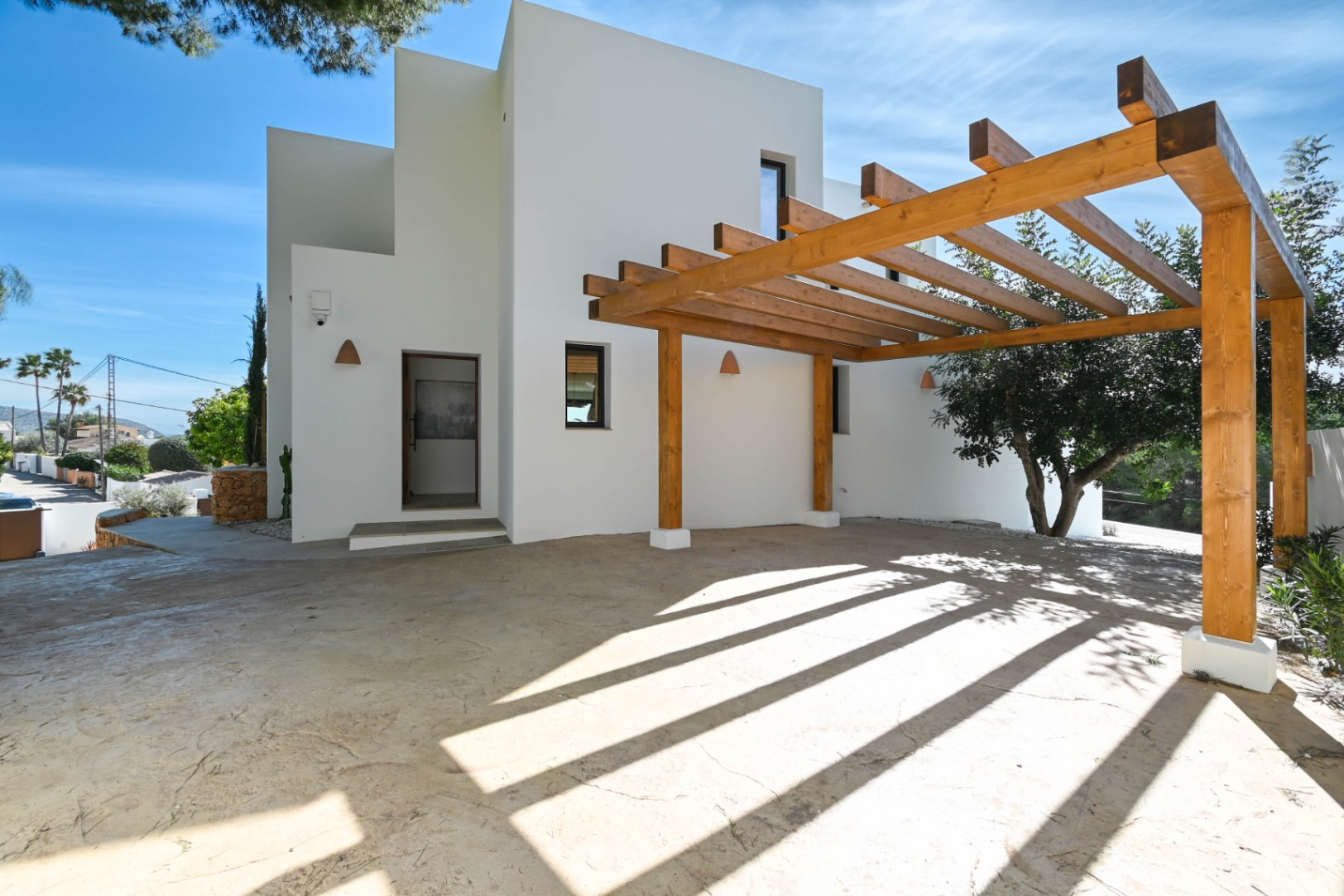 Minimalist white facade featuring a large natural wood beam pergola over a paved parking area. Includes ceramic wall sconces and a clean architectural entrance.
