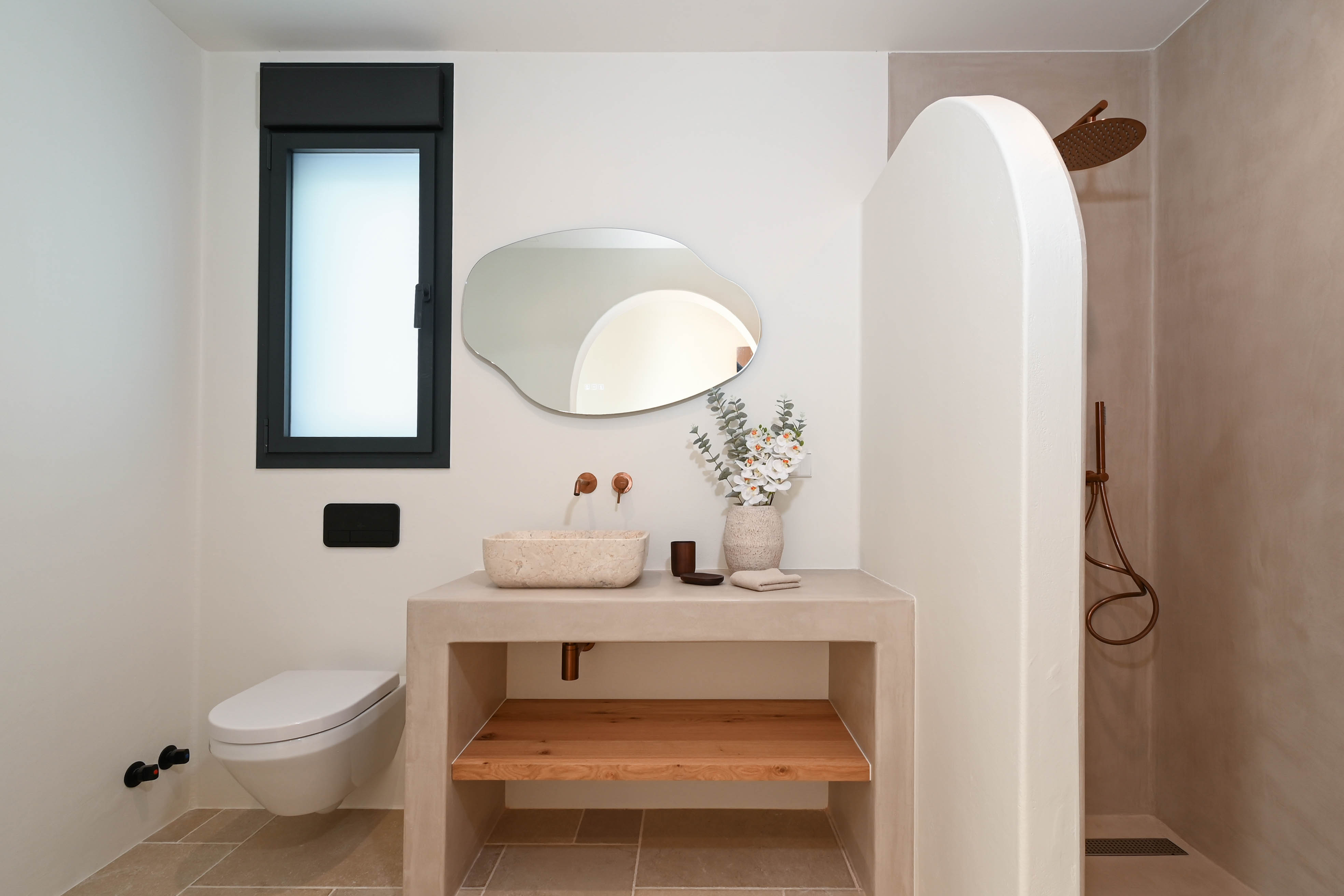 Modern bathroom featuring a microcement vanity, natural stone basin, and copper fixtures. It includes an organic-shaped mirror, wall-hung toilet, and a walk-in shower with a curved wall.