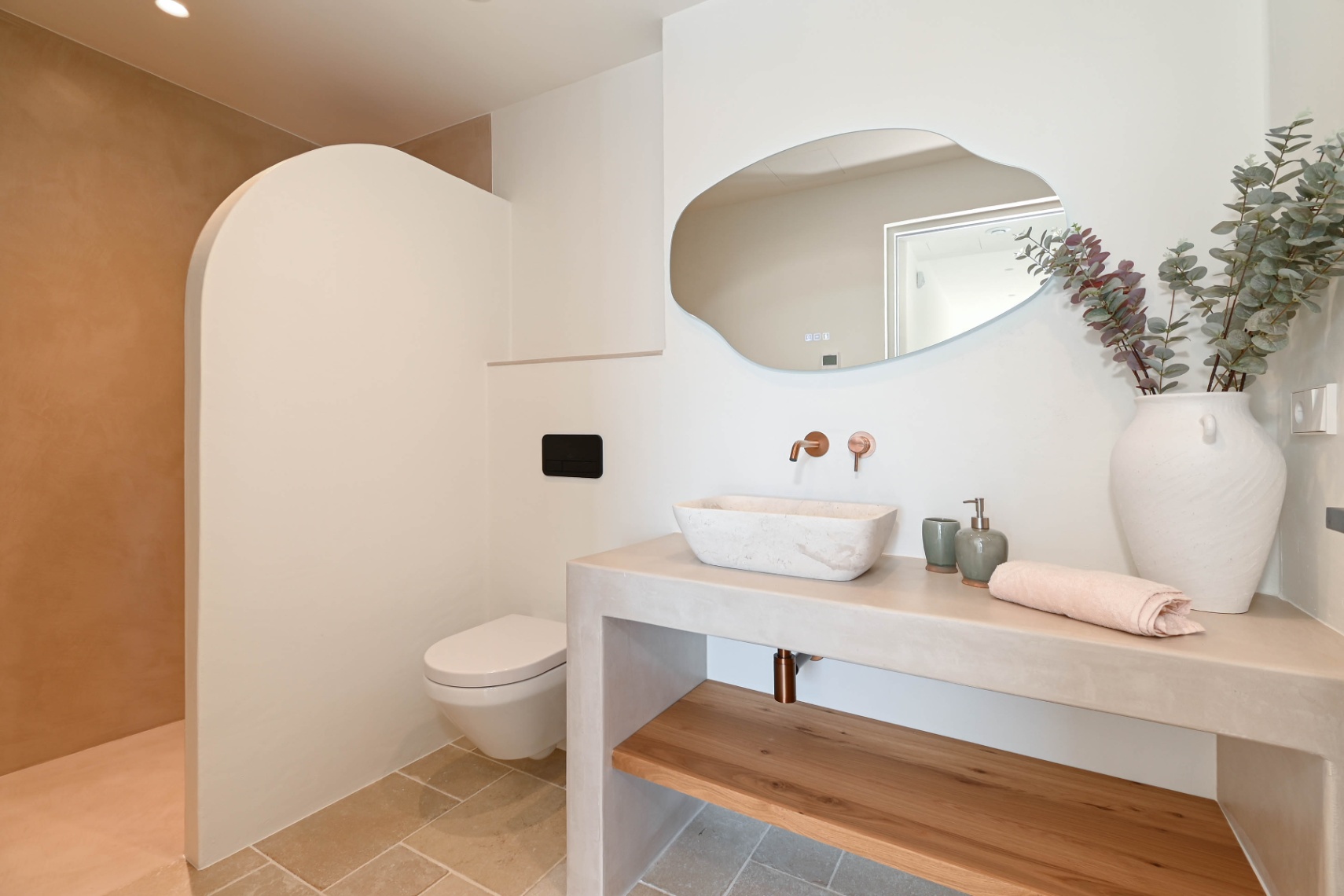 Modern bathroom featuring a stone vessel sink on a microcement vanity, copper wall-mounted fixtures, an organic-shaped mirror, and a curved shower partition wall.