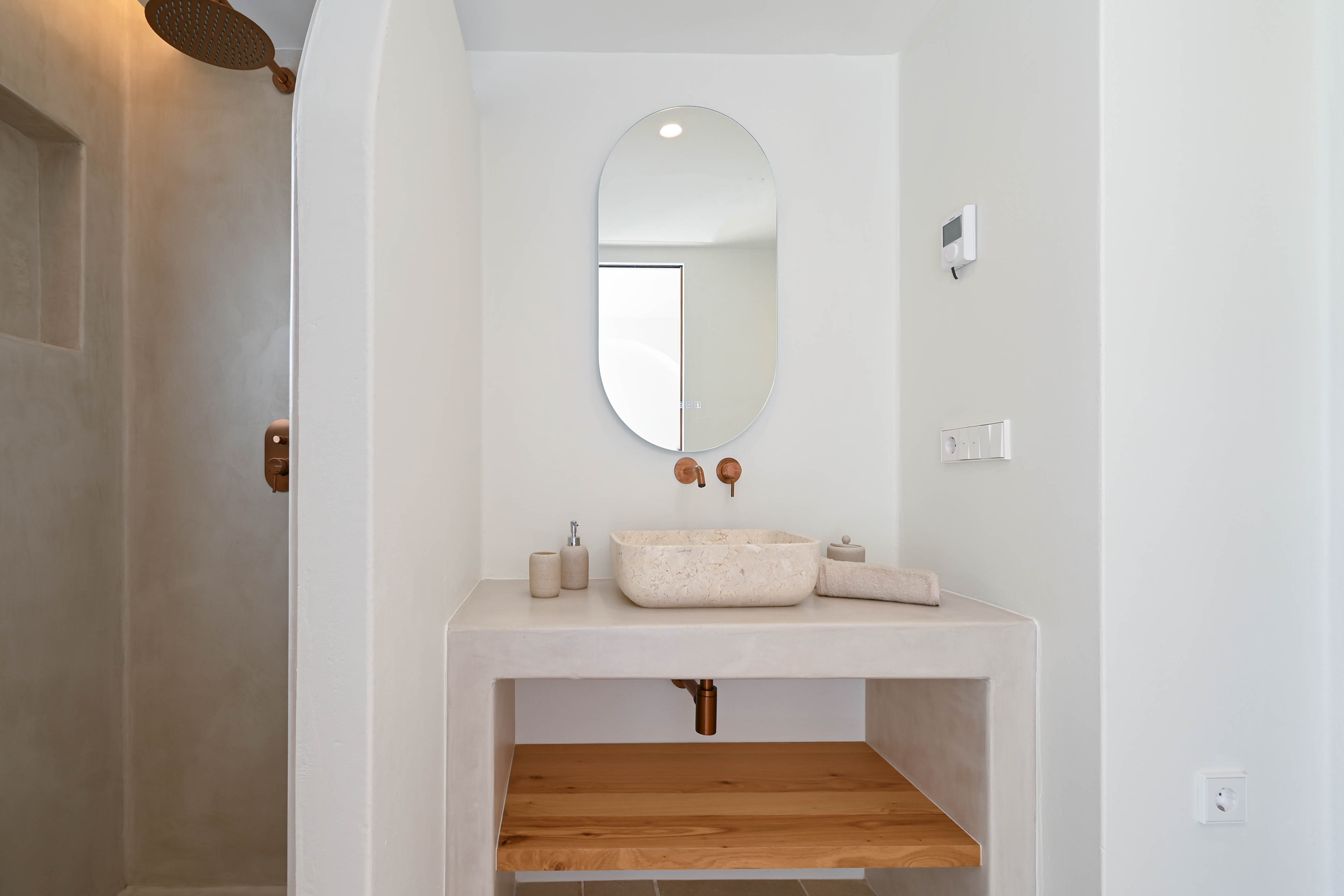 Modern bathroom featuring microcement finishes, a natural stone vessel sink, and copper hardware. A built-in vanity with an oak shelf and walk-in shower complete the space.