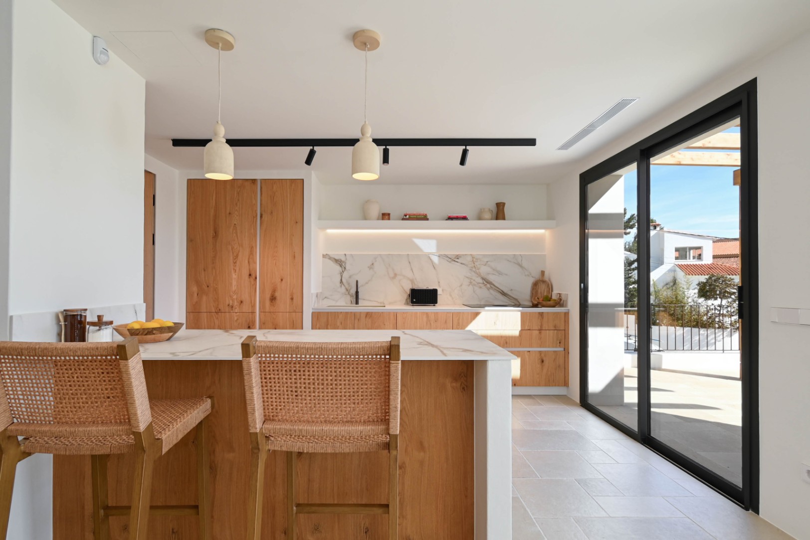 Open-concept kitchen featuring a wood island, marble-effect countertops, and woven stools, seamlessly connected to the terrace via large black-framed sliding doors.