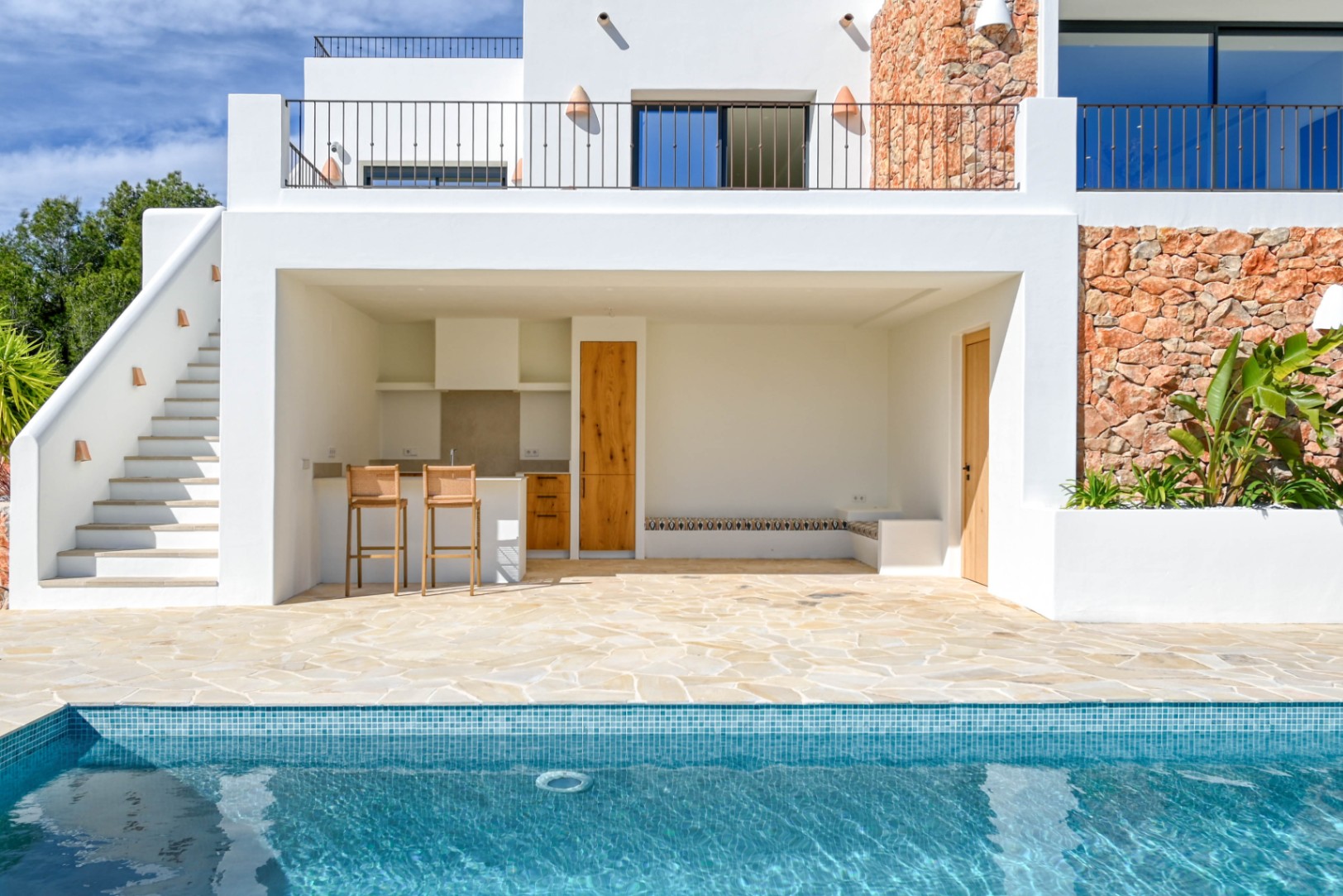 Outdoor kitchen and lounge area adjacent to the pool, featuring natural stone wall cladding, wooden cabinetry, and a stone-paved terrace under a white facade.