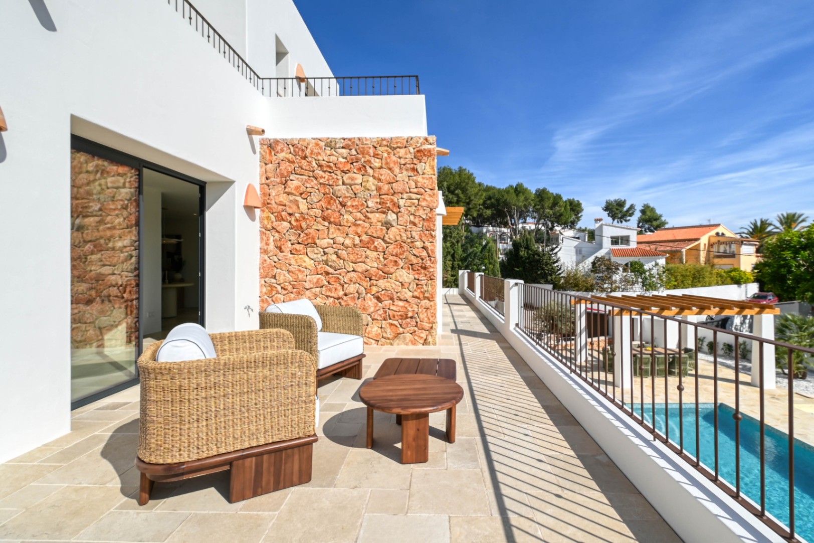 Upper terrace with natural stone tiling, wicker seating, and a dry-stone feature wall, offering views of the swimming pool and the lower pergola dining area.