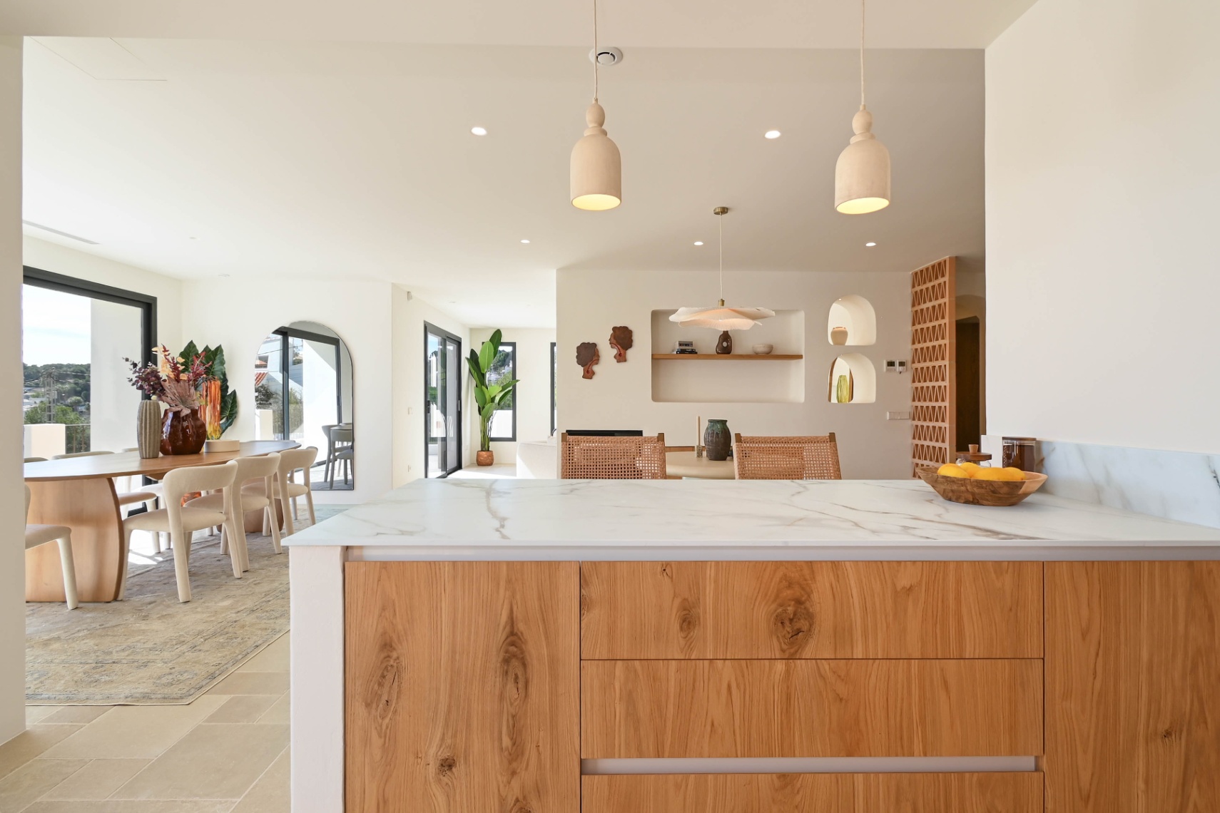 View from the veined stone kitchen island towards the open-plan dining area, featuring recessed wall niches, natural wood cabinetry, and large sliding glass doors.