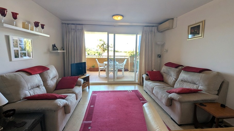 Bright living room featuring neutral tiled floors, dual sofas with red accents, and sliding glass doors opening onto a furnished terrace with garden views.