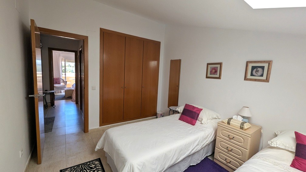 Bright twin bedroom with built-in wooden wardrobes and tiled floors. The open doorway reveals a hallway leading toward a sunlit room with terrace access.