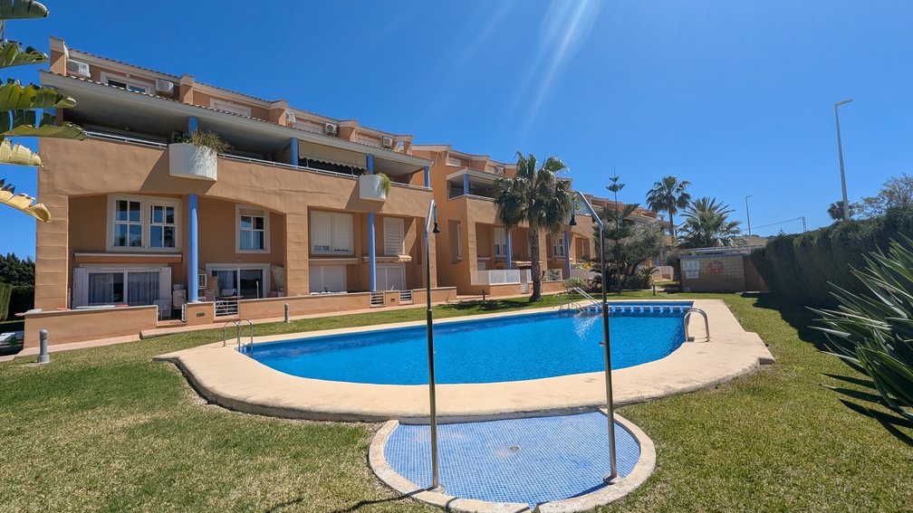 Communal kidney-shaped swimming pool surrounded by a manicured lawn and Mediterranean gardens, with the terracotta-colored apartment building and blue pillars in the background.