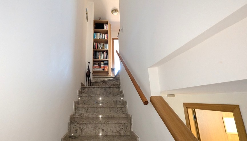 Internal marble staircase with a wooden handrail leading to the upper level, featuring a recessed bookshelf and clean white walls.
