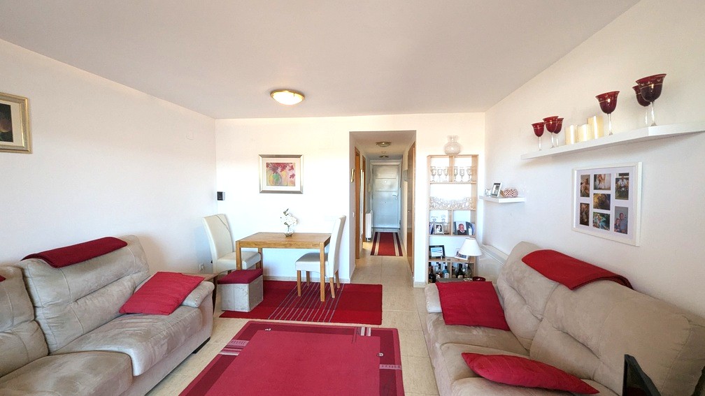 Living-dining area with beige sofas, red accents, and a wooden dining table. The layout connects to a central hallway with light tile flooring and white walls.