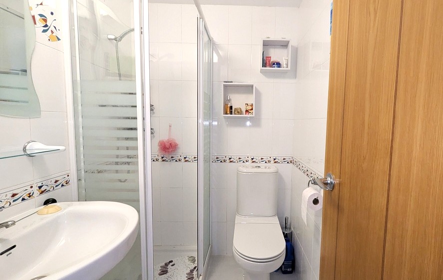 Modern shower room featuring white wall tiles with a decorative border, a glass-enclosed shower unit, and integrated shelving for storage.