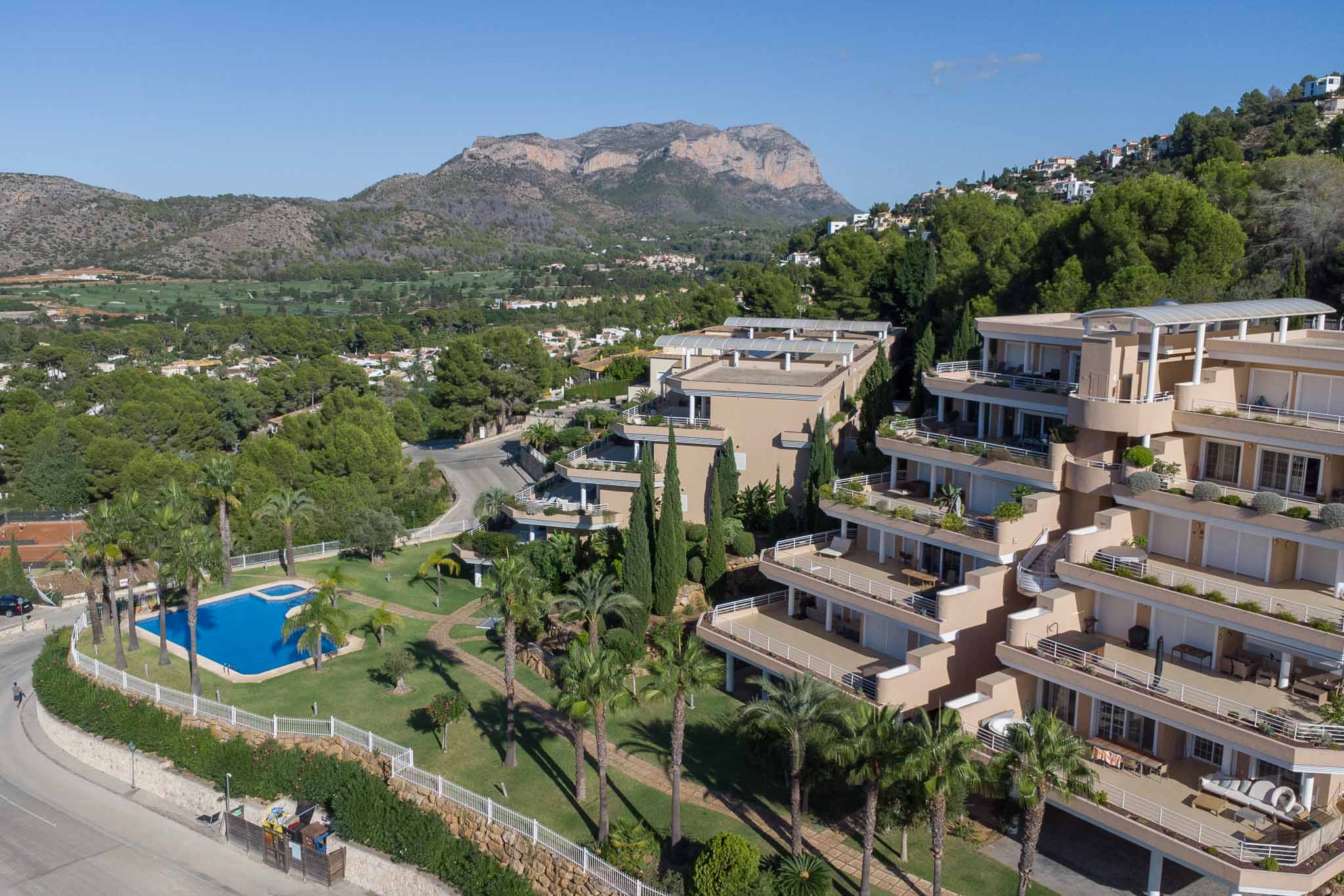 Aerial perspective of the Las Mimosas residential complex, showcasing the communal pool, manicured lawns, and terraced buildings set against the Montgó mountain.