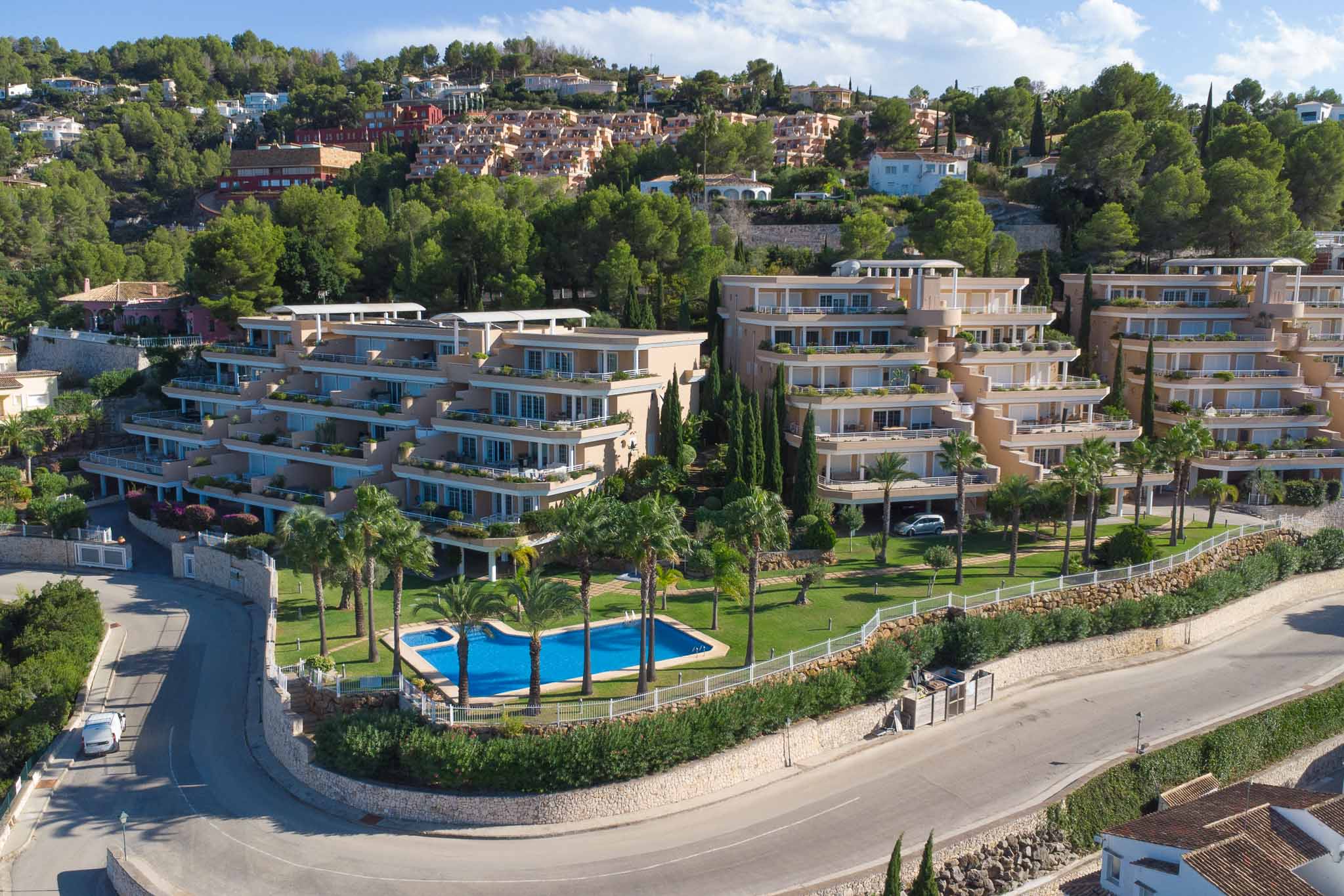 Aerial view of the Las Mimosas complex in La Sella, featuring terraced architecture, a communal swimming pool with palm trees, and manicured gardens.