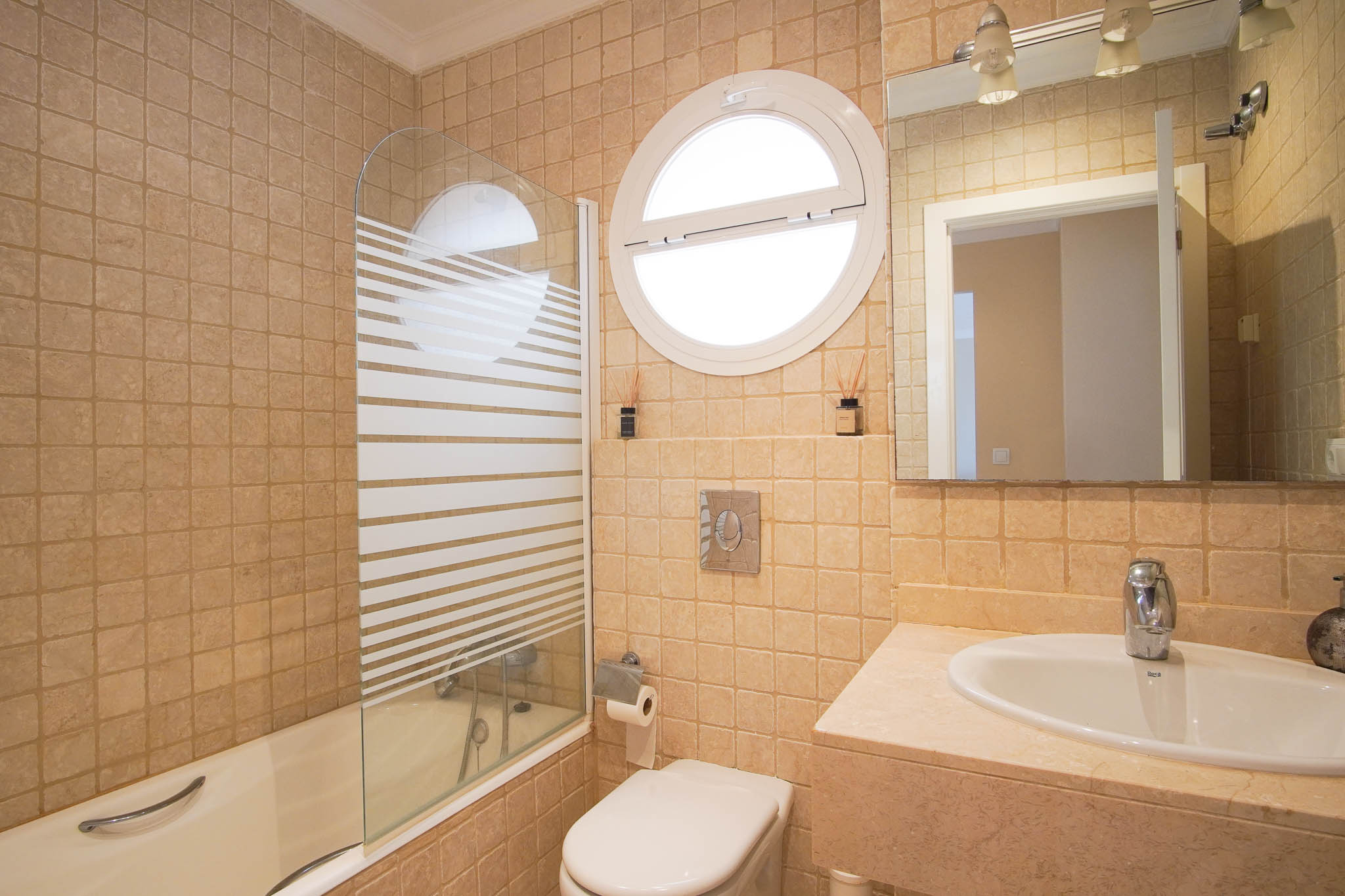 Bathroom featuring marble surfaces, a bathtub with a striped glass screen, and a unique circular window providing natural light.