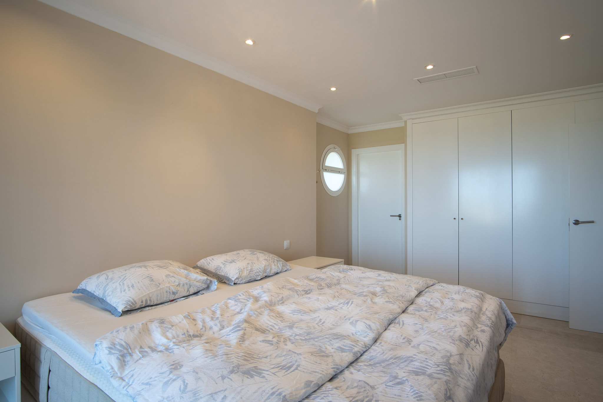 Bedroom featuring a double bed, floor-to-ceiling white wardrobes, and marble flooring. The space is lit by recessed ceiling spots and a unique porthole window.