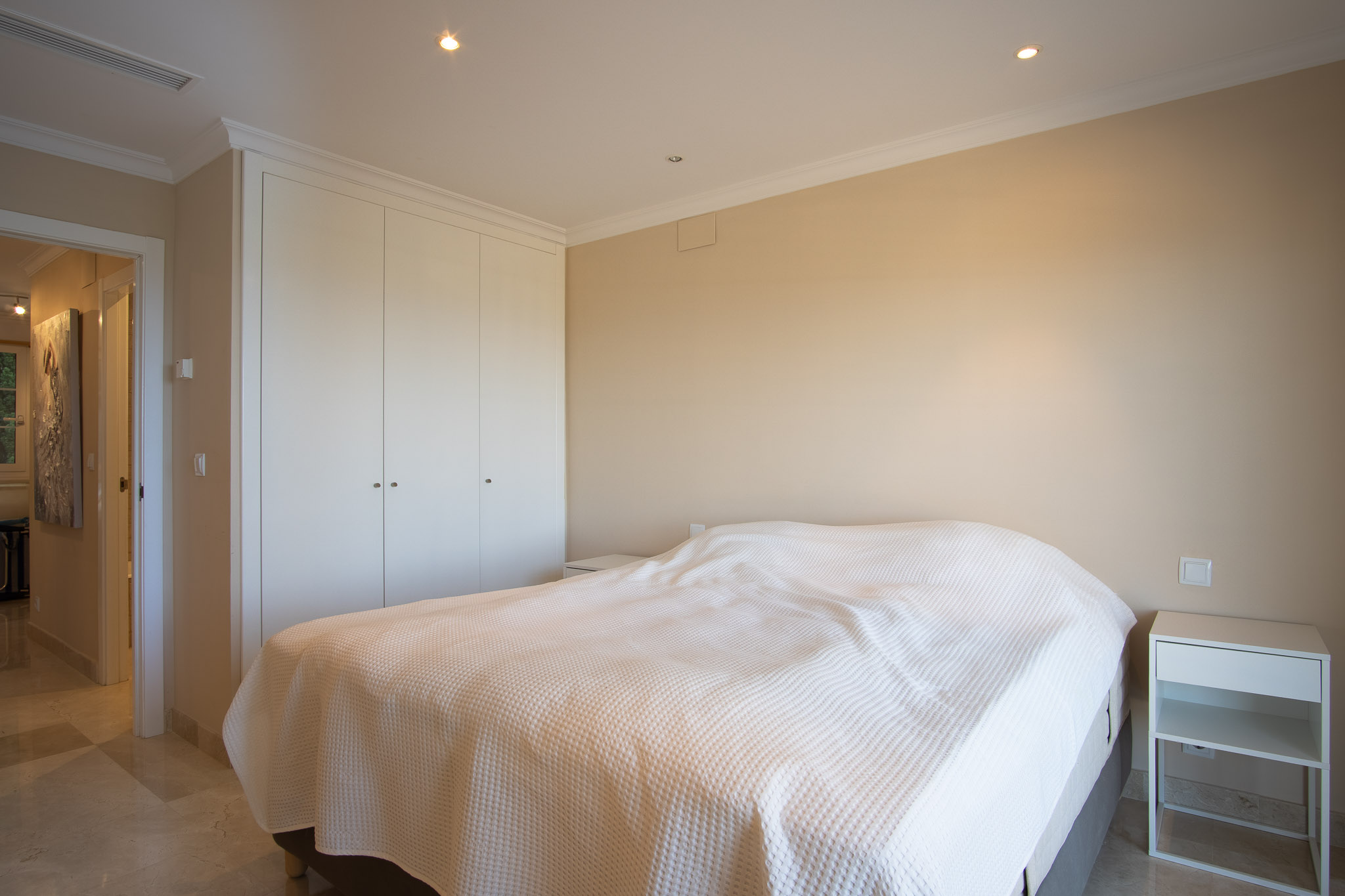 Bedroom featuring marble flooring, floor-to-ceiling white built-in wardrobes, and recessed ceiling spotlights, showcasing a clean and functional interior design.
