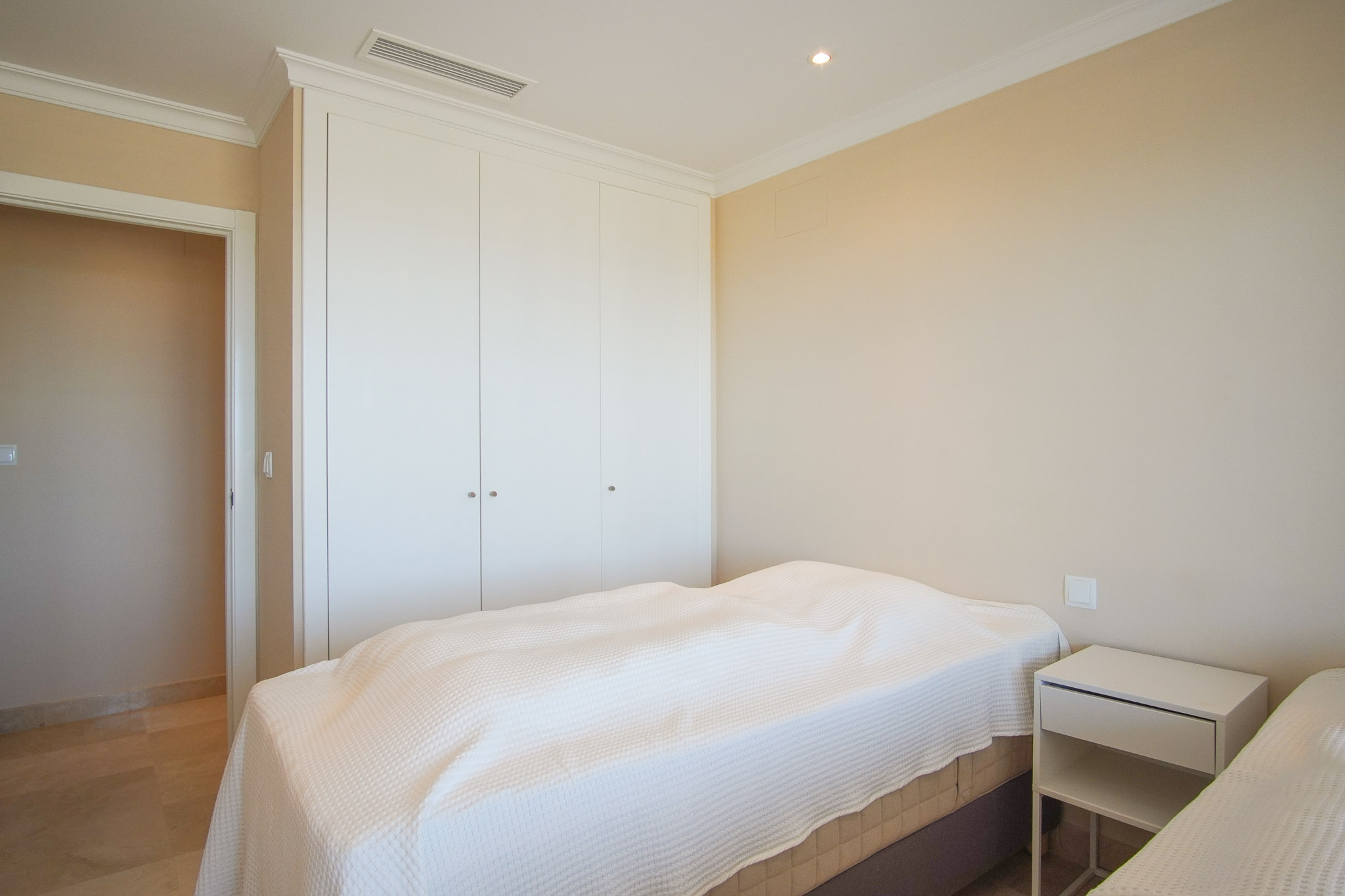Bright bedroom featuring floor-to-ceiling white wardrobes, marble flooring, and ducted air conditioning. Neutral cream walls and elegant crown molding.