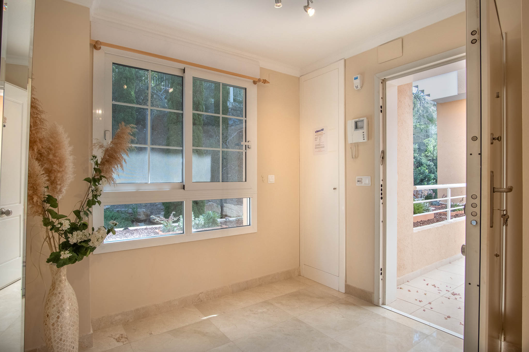 Bright entrance hall featuring polished marble flooring, a double-glazed window overlooking greenery, and a secure main entry door.