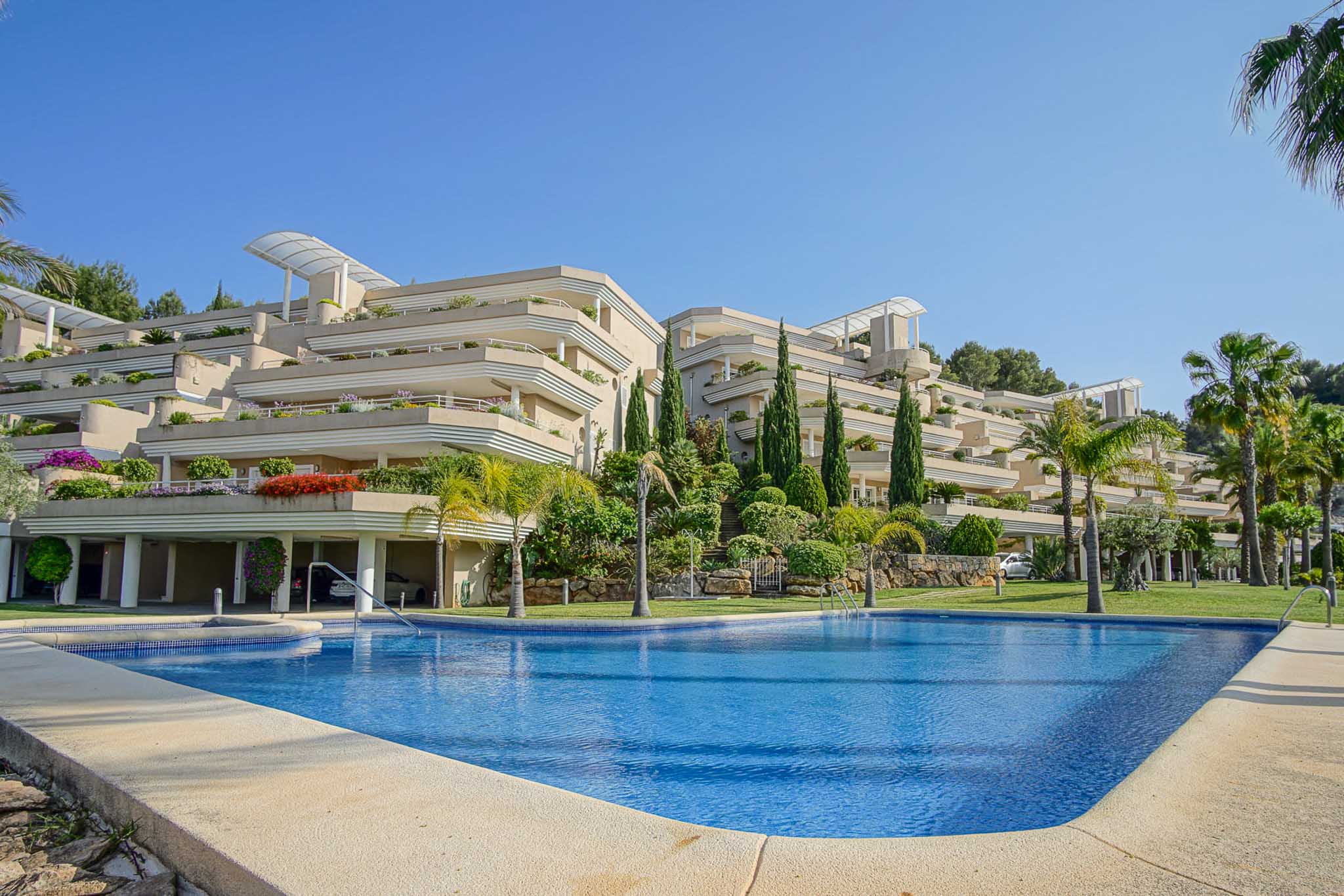Exterior view of the Las Mimosas complex, featuring a large communal pool and terraced architecture with integrated gardens and palm trees under a clear sky.