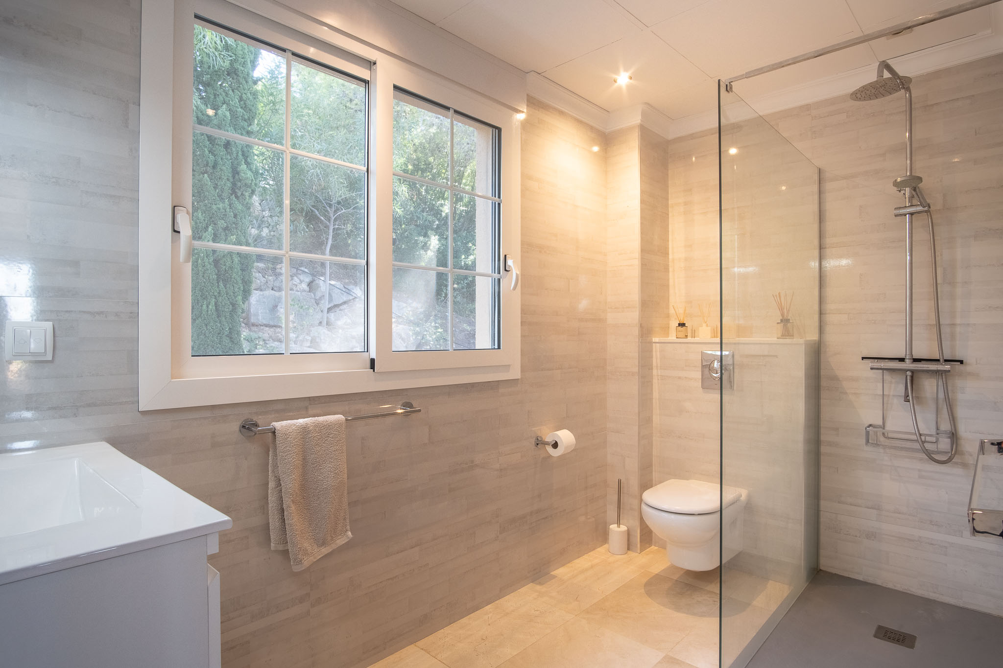 Modern bathroom featuring a walk-in shower with glass partition and rain shower head. It includes wall-mounted fixtures, neutral tiling, and a large window providing natural light.