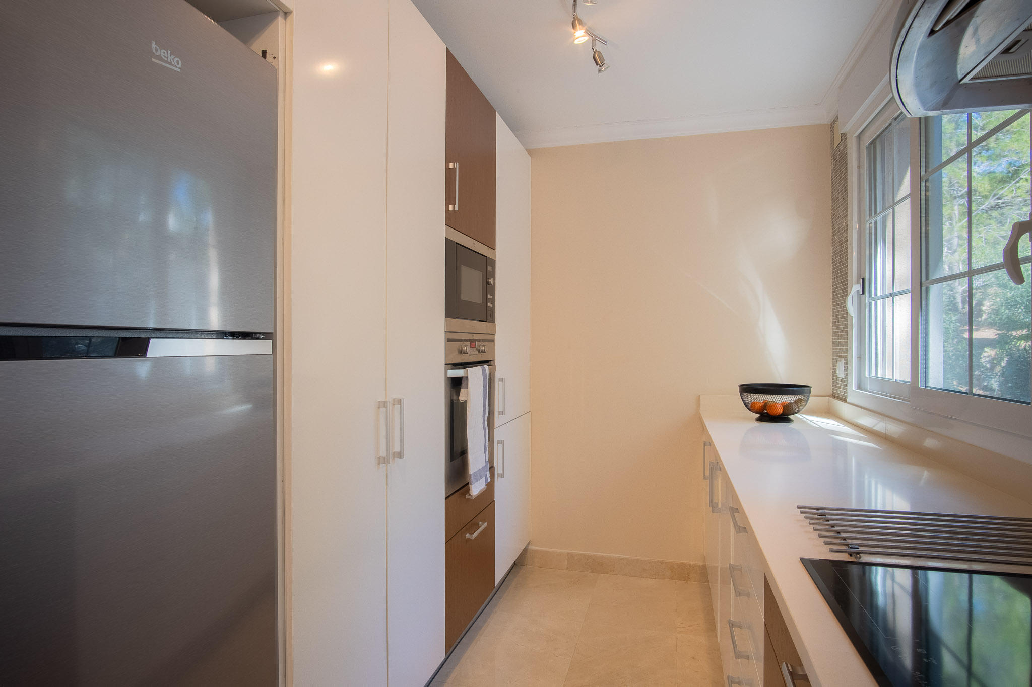 Modern kitchen featuring white and wood-finish cabinetry, integrated stainless steel appliances, and a white countertop. A side window provides natural light over the induction hob.