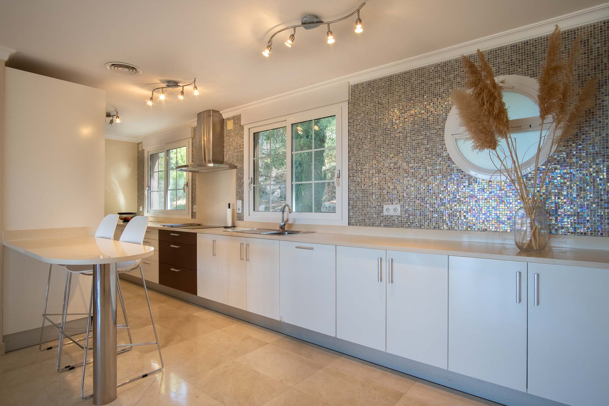 Modern kitchen featuring white cabinetry, a light countertop, and a distinctive iridescent mosaic backsplash. Includes a breakfast bar, marble flooring, and natural light.