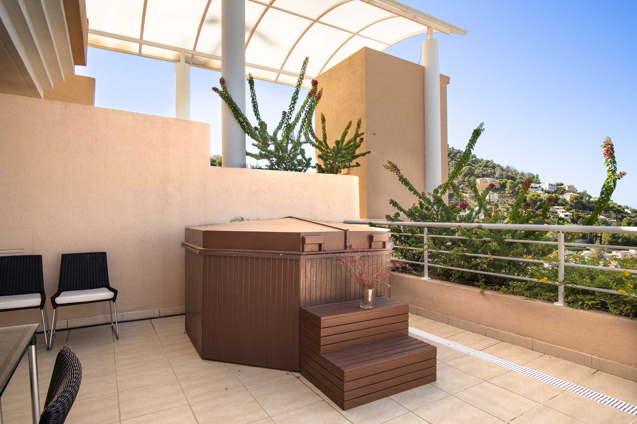 Private terrace featuring a covered jacuzzi with wooden steps, situated under an arched pergola with views of the surrounding hills and Mediterranean flora.