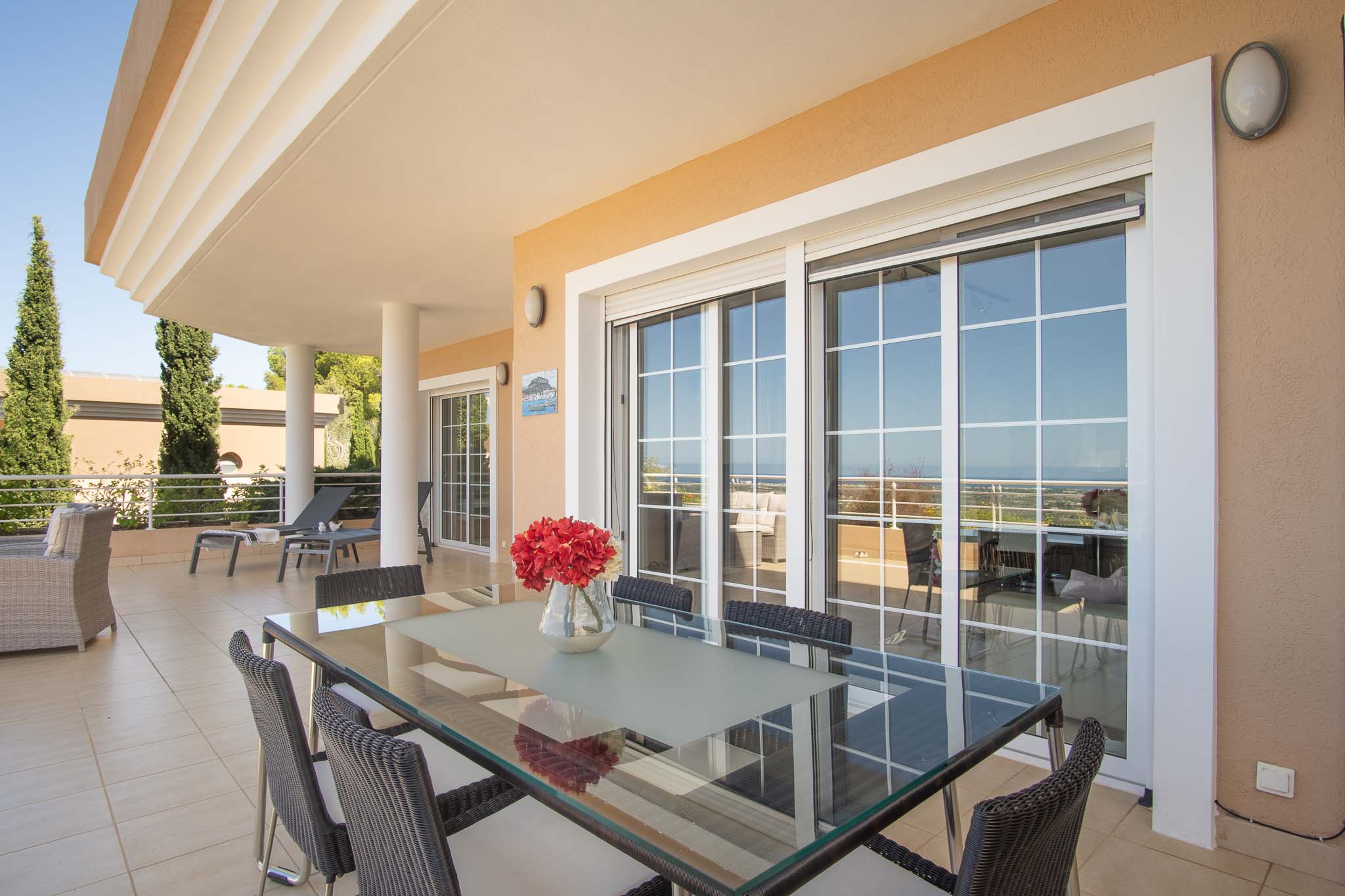 Spacious covered terrace featuring an outdoor dining area with a glass-top table, wicker chairs, and large sliding doors overlooking the Mediterranean coast.