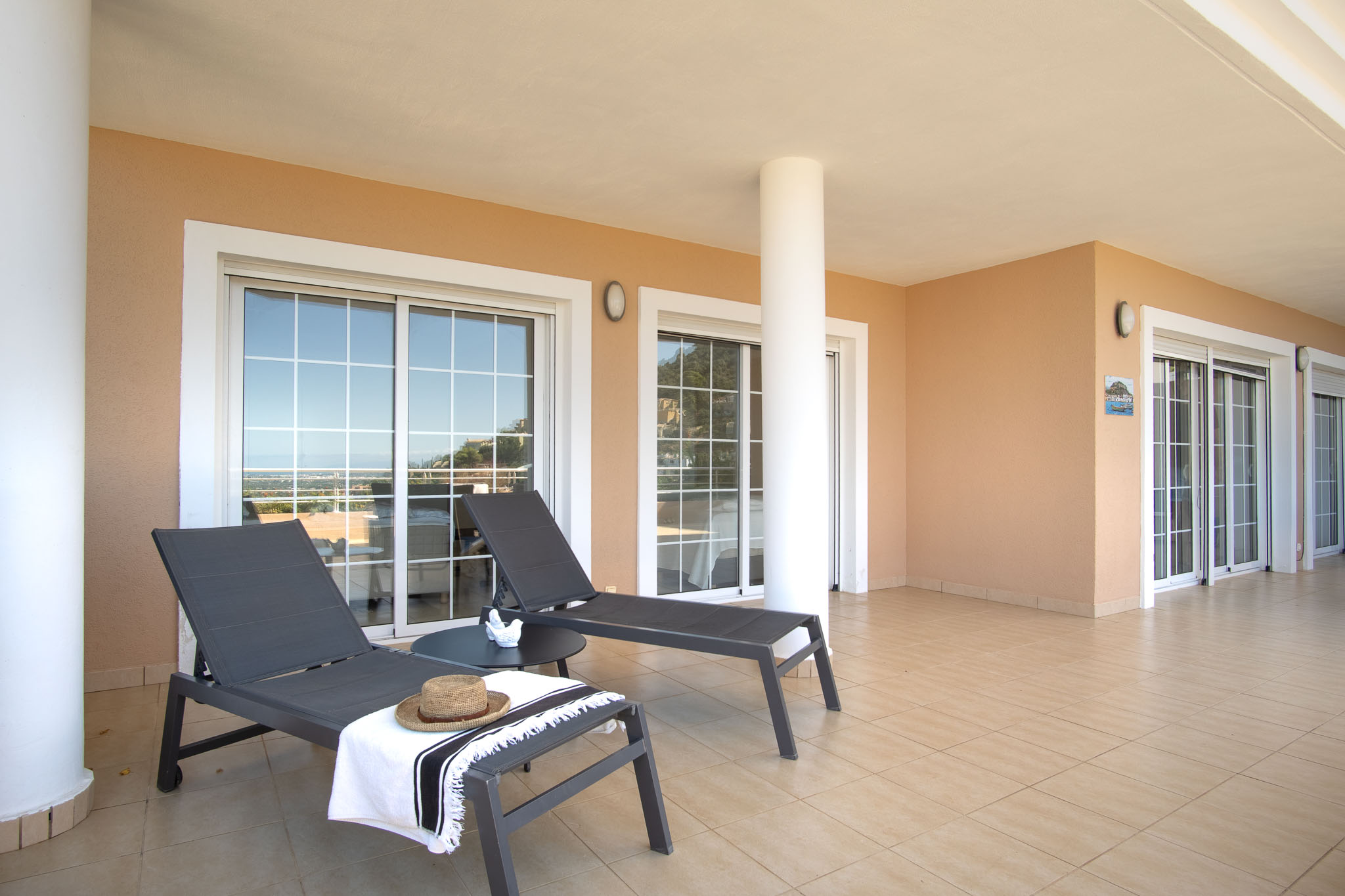 Spacious covered terrace featuring ceramic tiling, white structural columns, and modern sun loungers. Multiple sliding glass doors provide seamless access to the interior rooms.