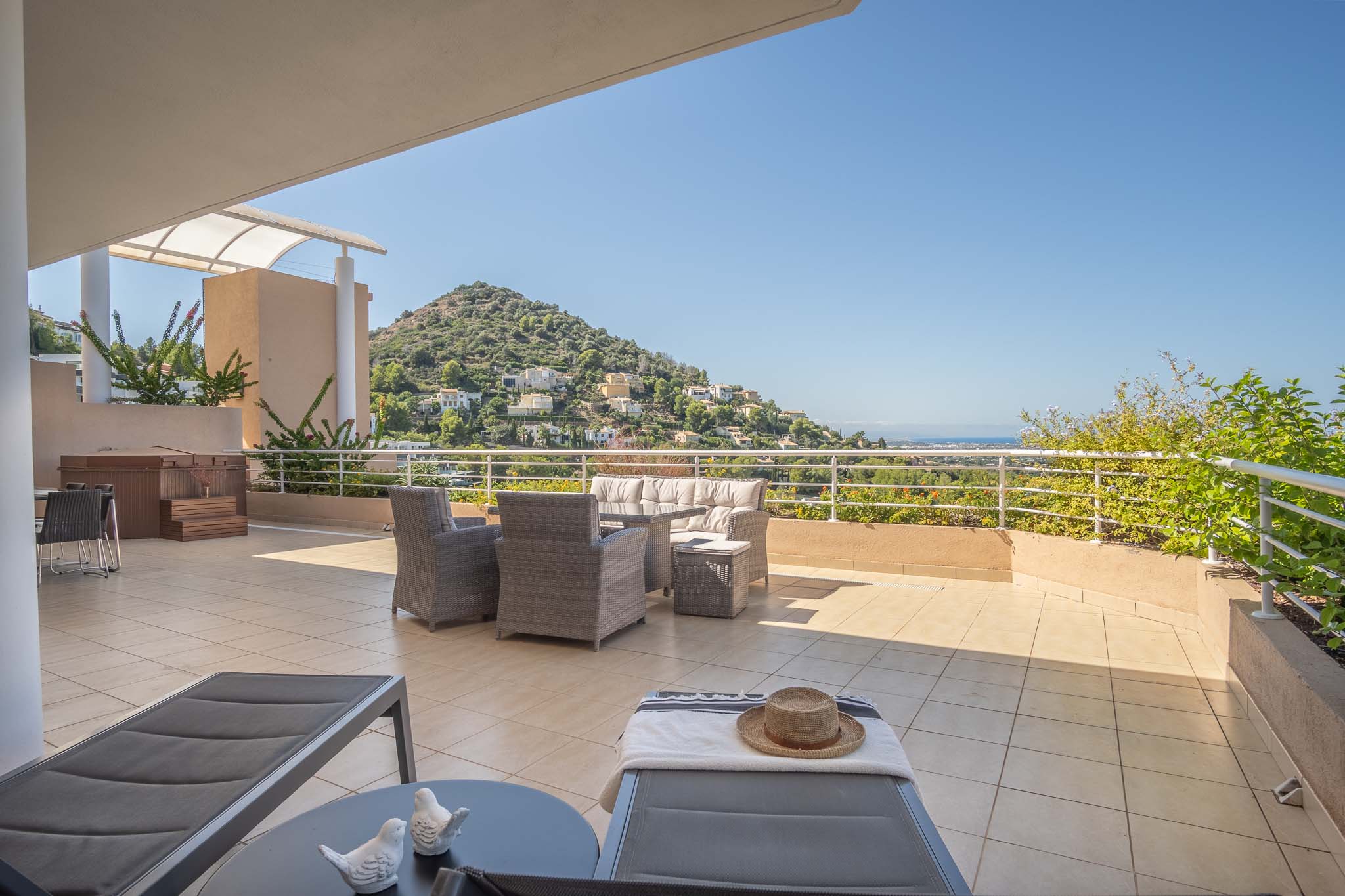 Spacious tiled terrace featuring a grey wicker lounge set, private jacuzzi, and sun loungers. It offers panoramic views of the surrounding hills and the Mediterranean Sea.