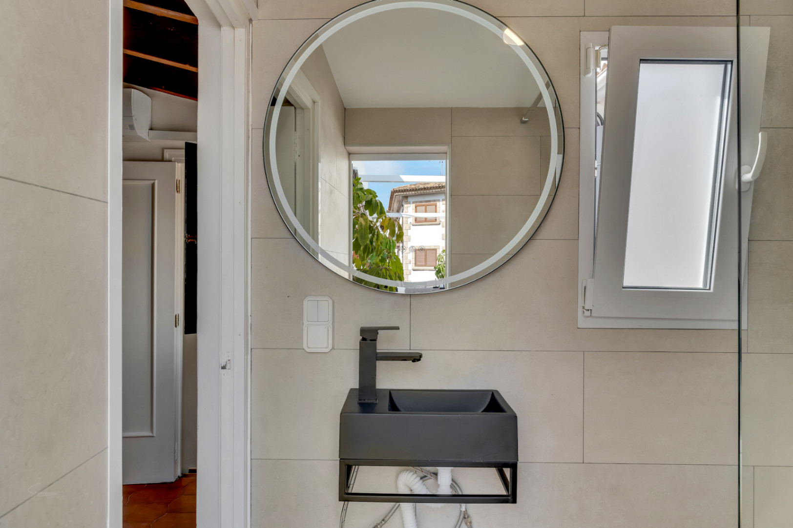 Bathroom detail featuring a black wall-mounted sink, round lighted mirror, and awning window.