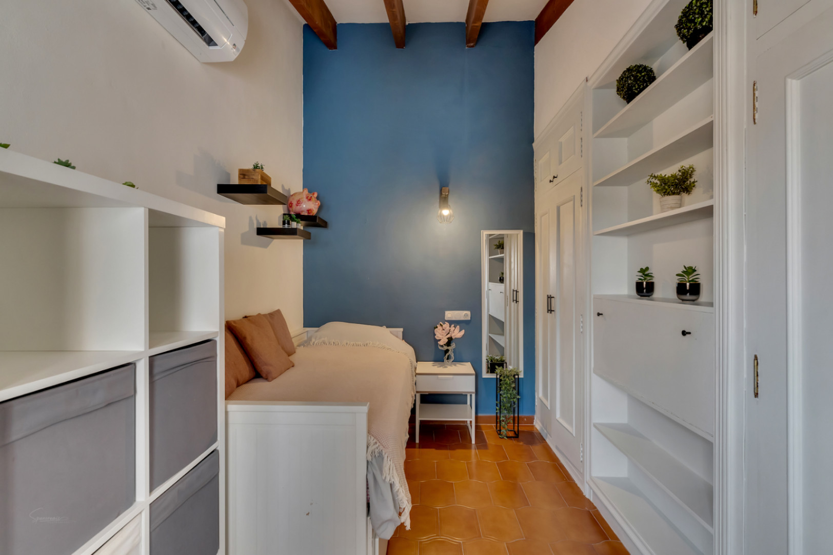 Bedroom with single bed, white shelving units, blue accent wall, and terracotta tile floor.