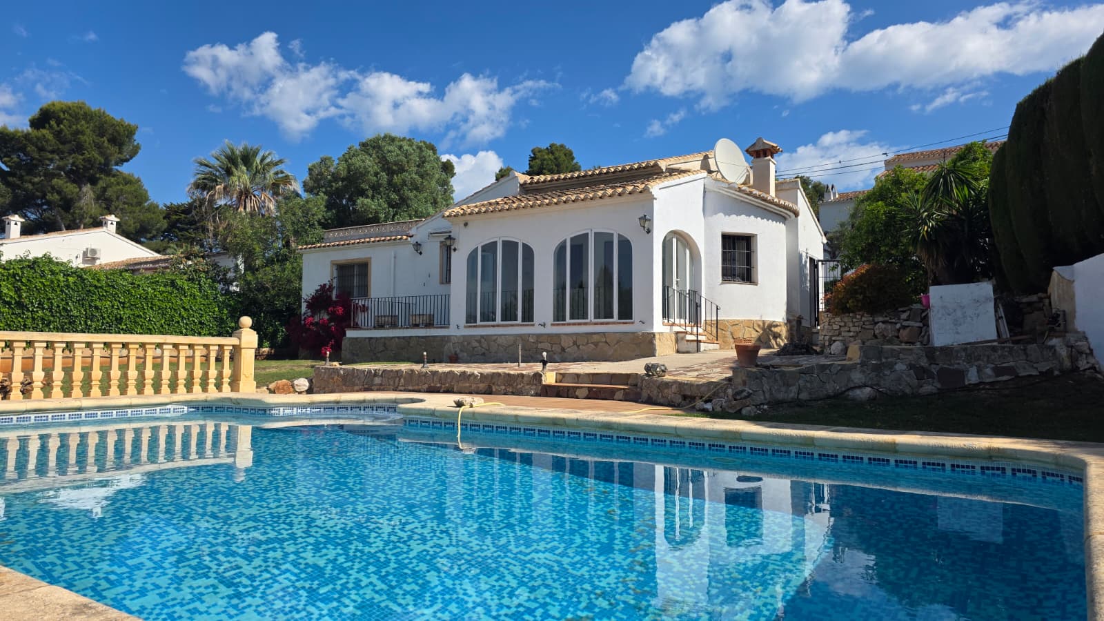 Exterior view of the private swimming pool and the white-walled villa with traditional arched windows and stone masonry base in El Tosalet, Jávea.