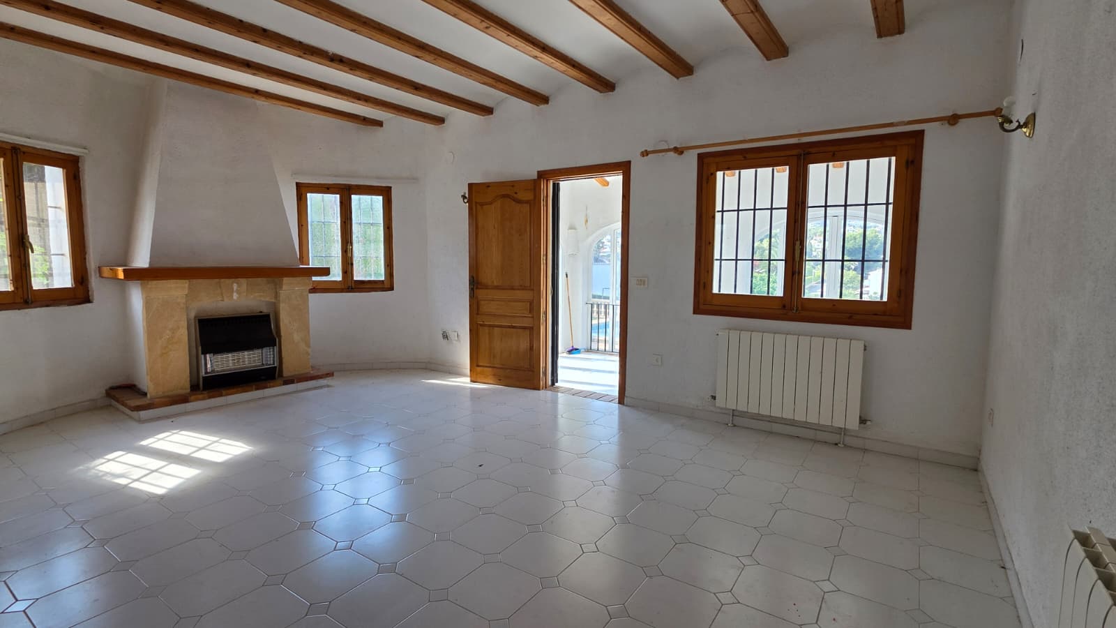 Living room featuring exposed wooden ceiling beams, a traditional stone fireplace, and white tiled flooring. Large wooden windows provide natural light.