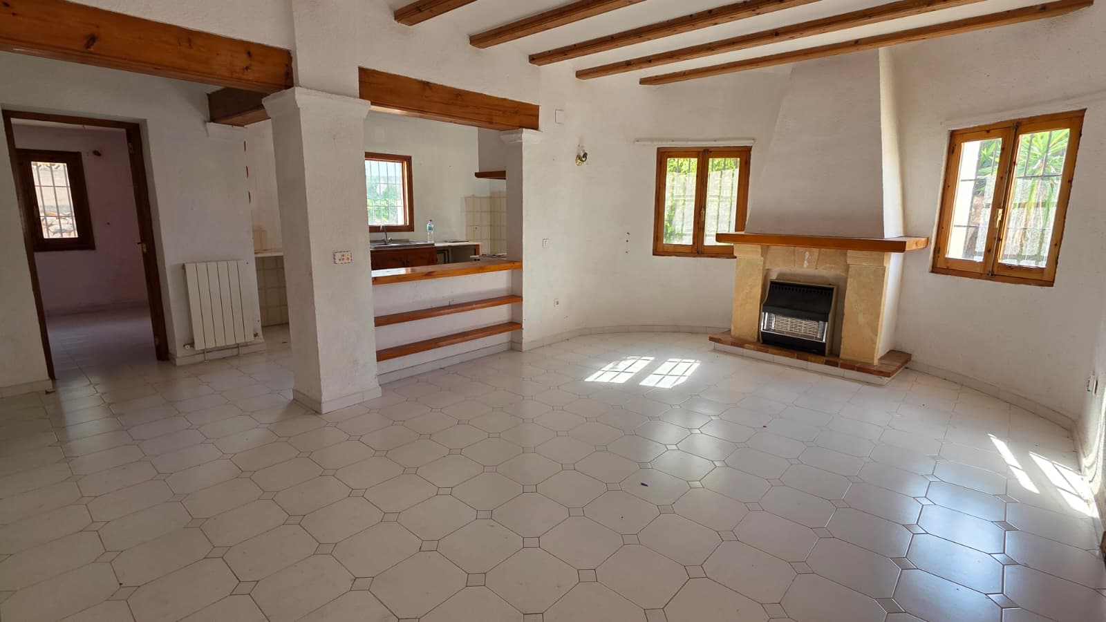 Spacious living area featuring traditional exposed wooden beams, a stone fireplace with integrated heater, and white tiled flooring. An open pass-through connects to the kitchen.