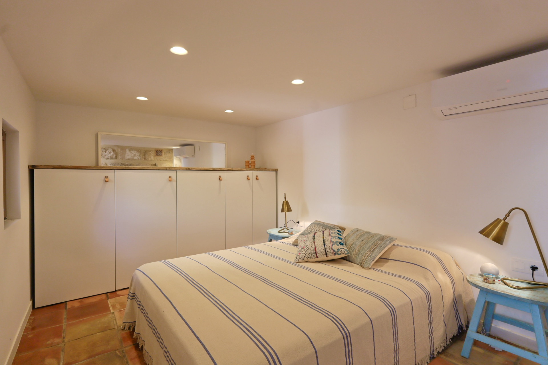 Bright bedroom with terracotta flooring, striped bedspread, and white built-in wardrobe with wood accents.